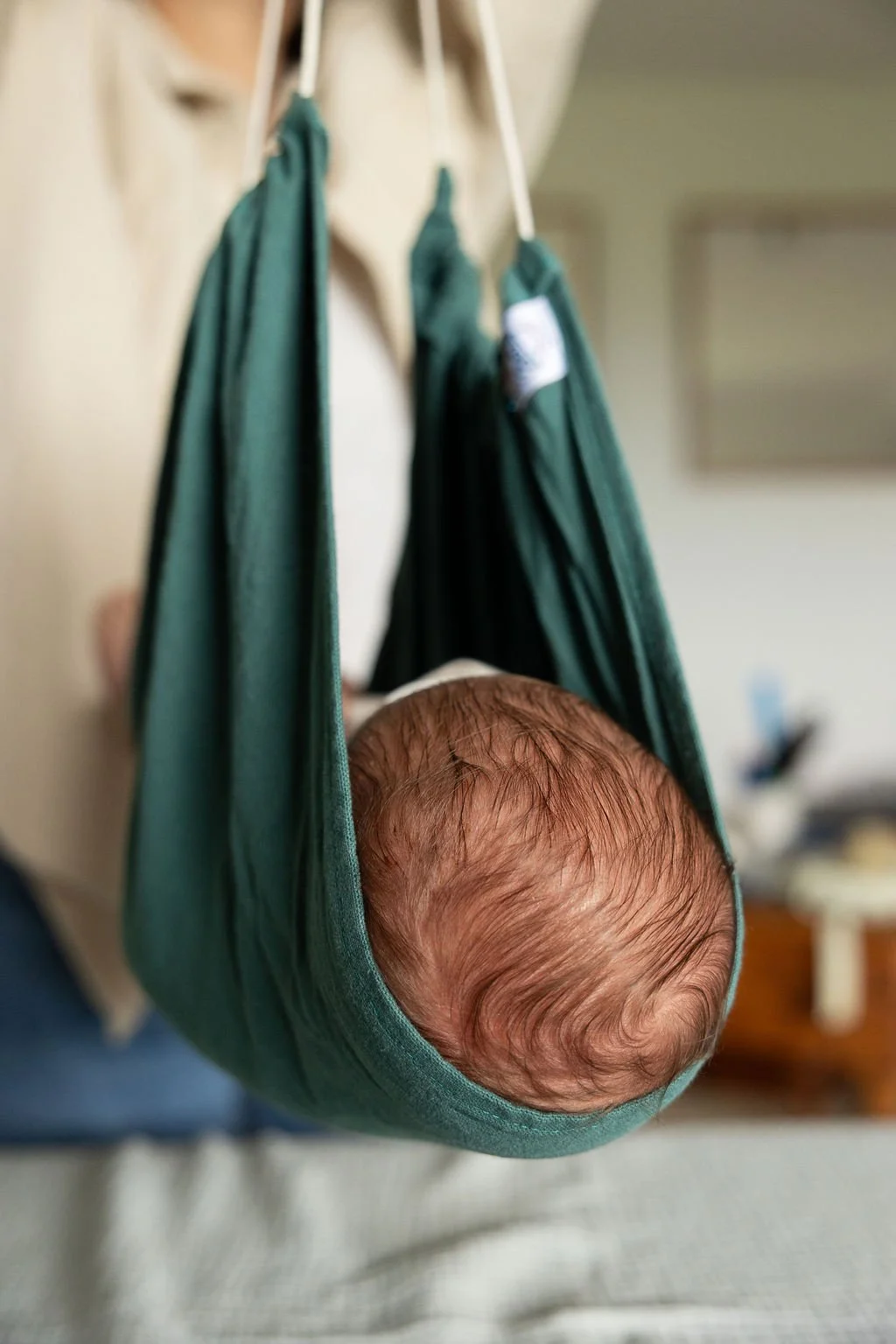Centrée sur la tête d'un nouveau-né avec des cheveux fins, dans un tissu vert suspendu à une structure en bois ou en métal, dans un intérieur domestique.