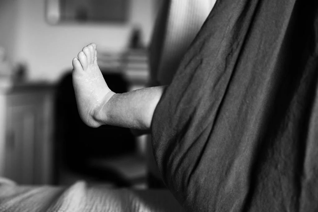 Un bébé repose ses jambes sur le genou d'un adulte dans un intérieur domestique, en noir et blanc.