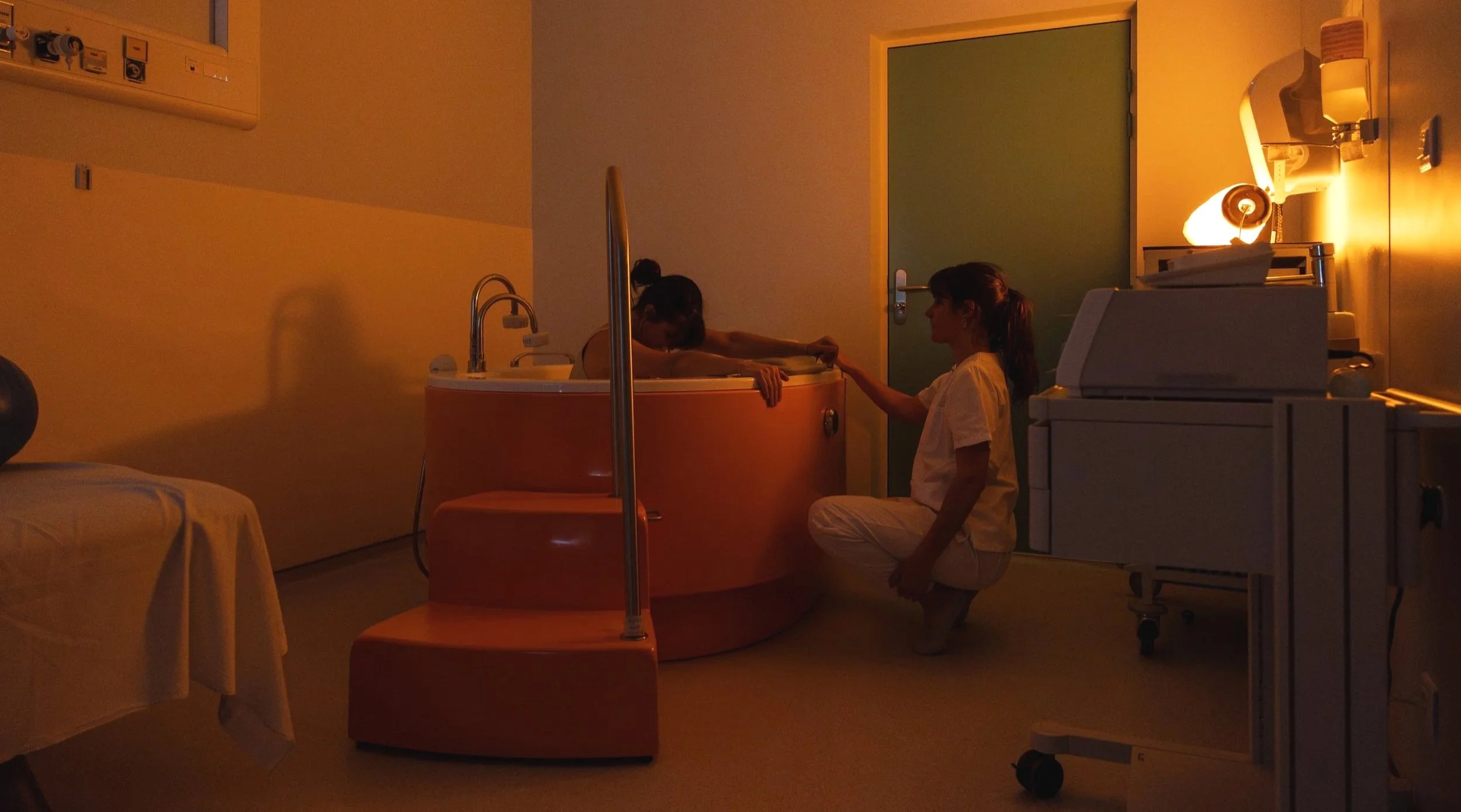 Deux personnes dans une salle de bain, une dans un bain de créme et l'autre assise à côté, dans une ambiance tamisée.