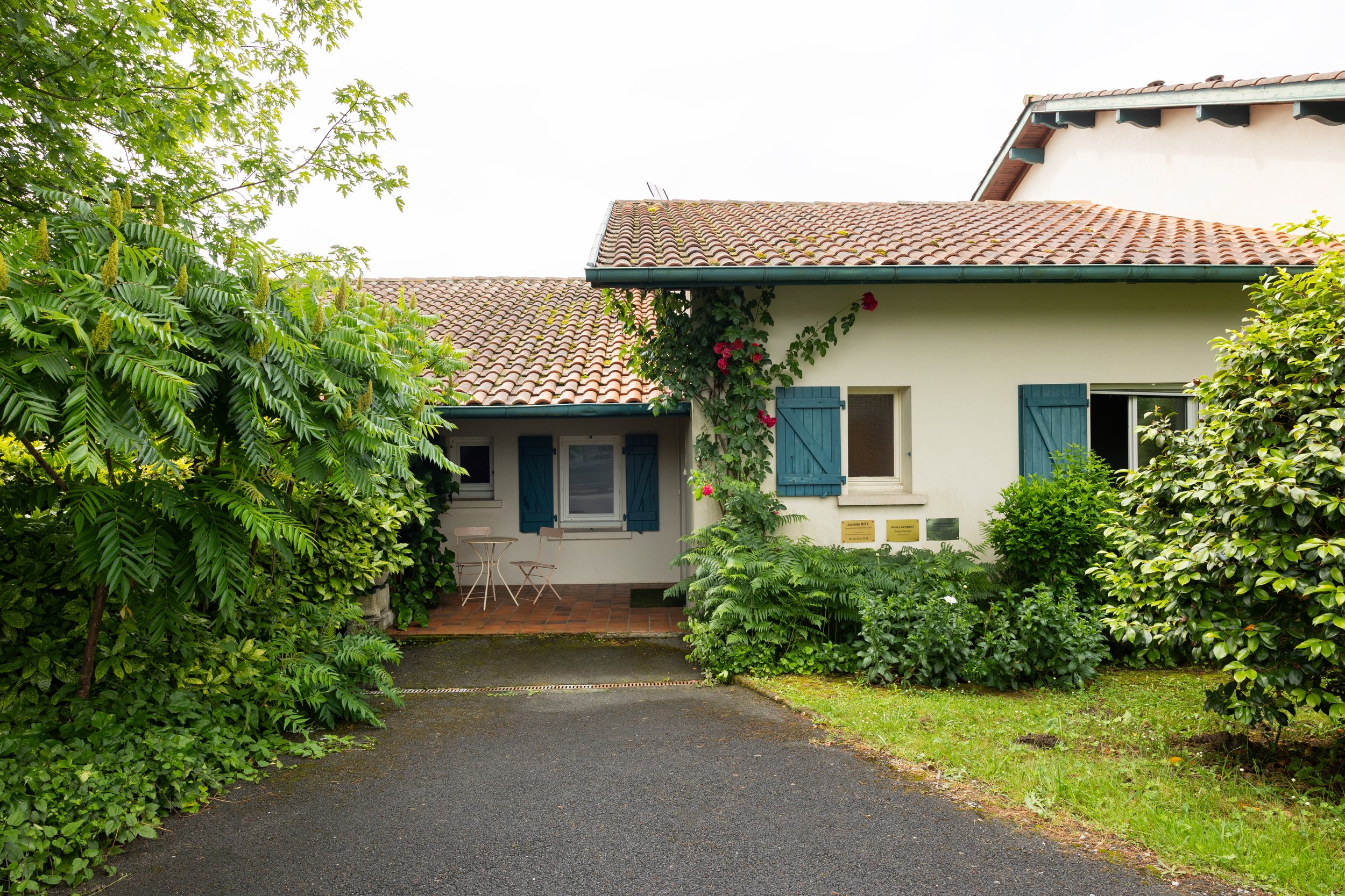 Maison blanche avec volets bleus, entourée de verdure, avec une petite terrasse et un toit en tuiles rouges.
