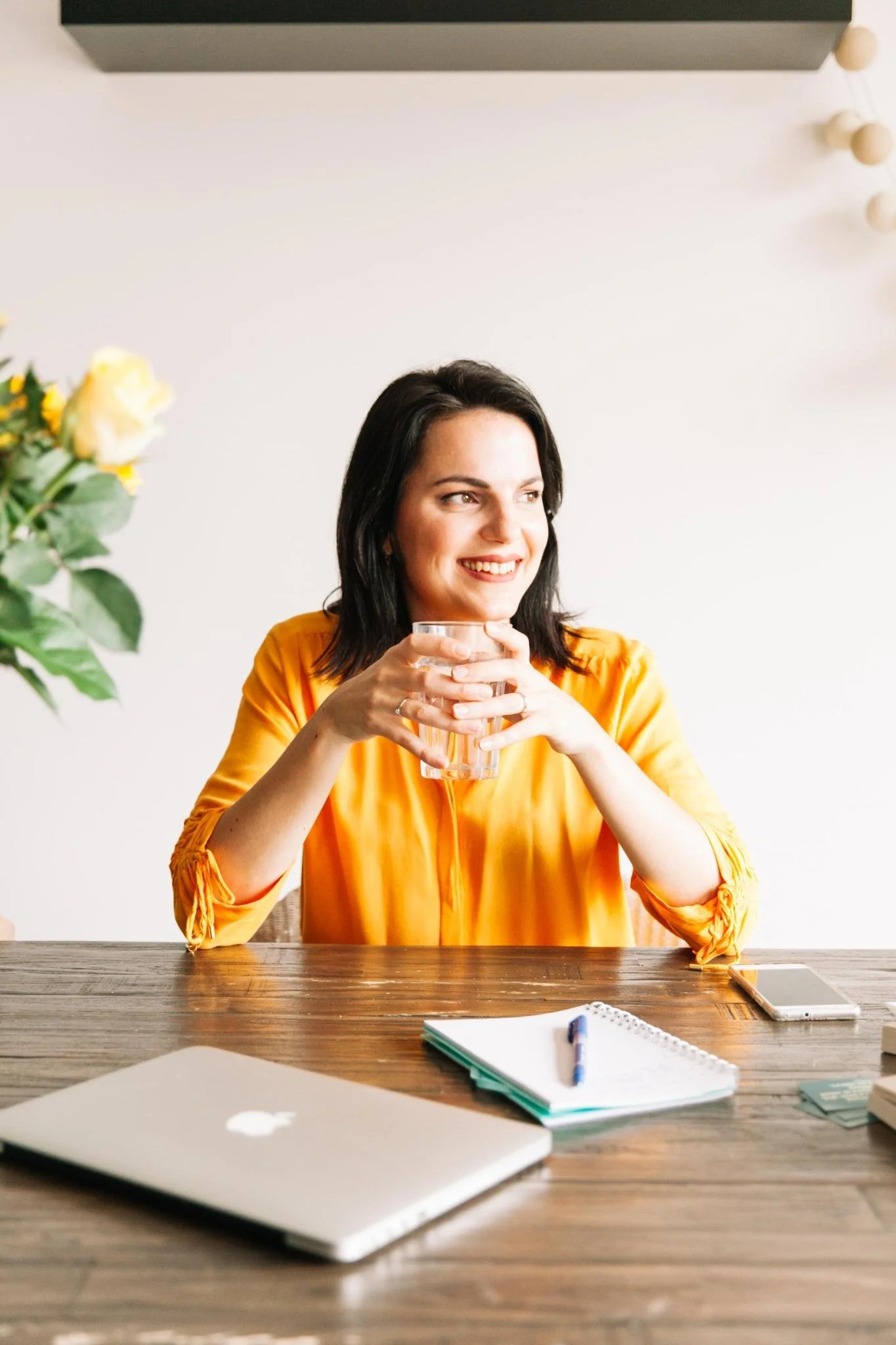 Vrouw met donkere haar in een gele blouse, lachend en een glas water vasthoudend, zittend aan een houten tafel met een laptop, notitieboekje, pen en smartphone.