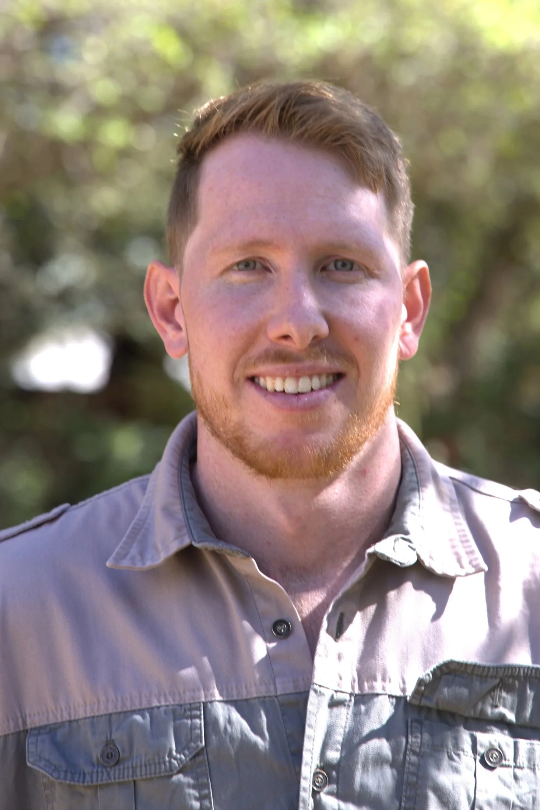Close-up of a young man with red hair and a light beard smiling outdoors, wearing a light gray button-up shirt.