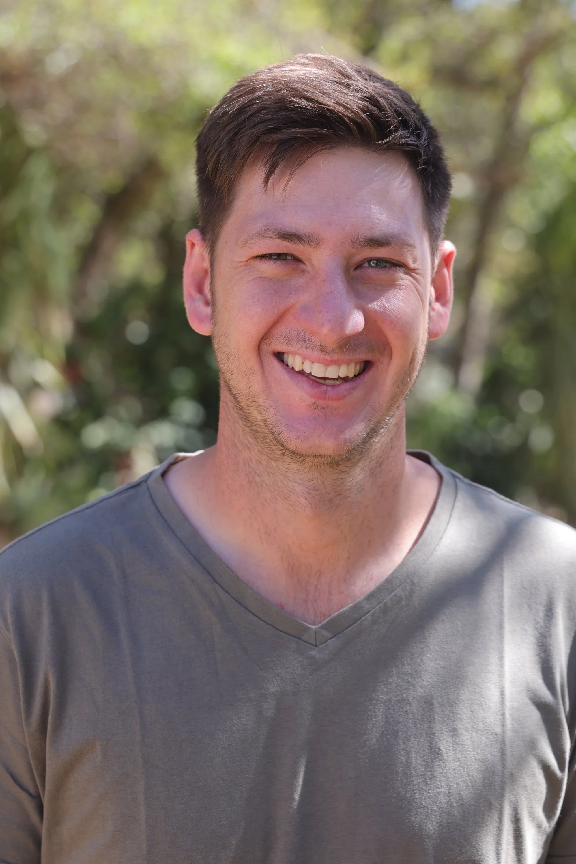 Close-up of a young man smiling outdoors with blurred green trees in the background.
