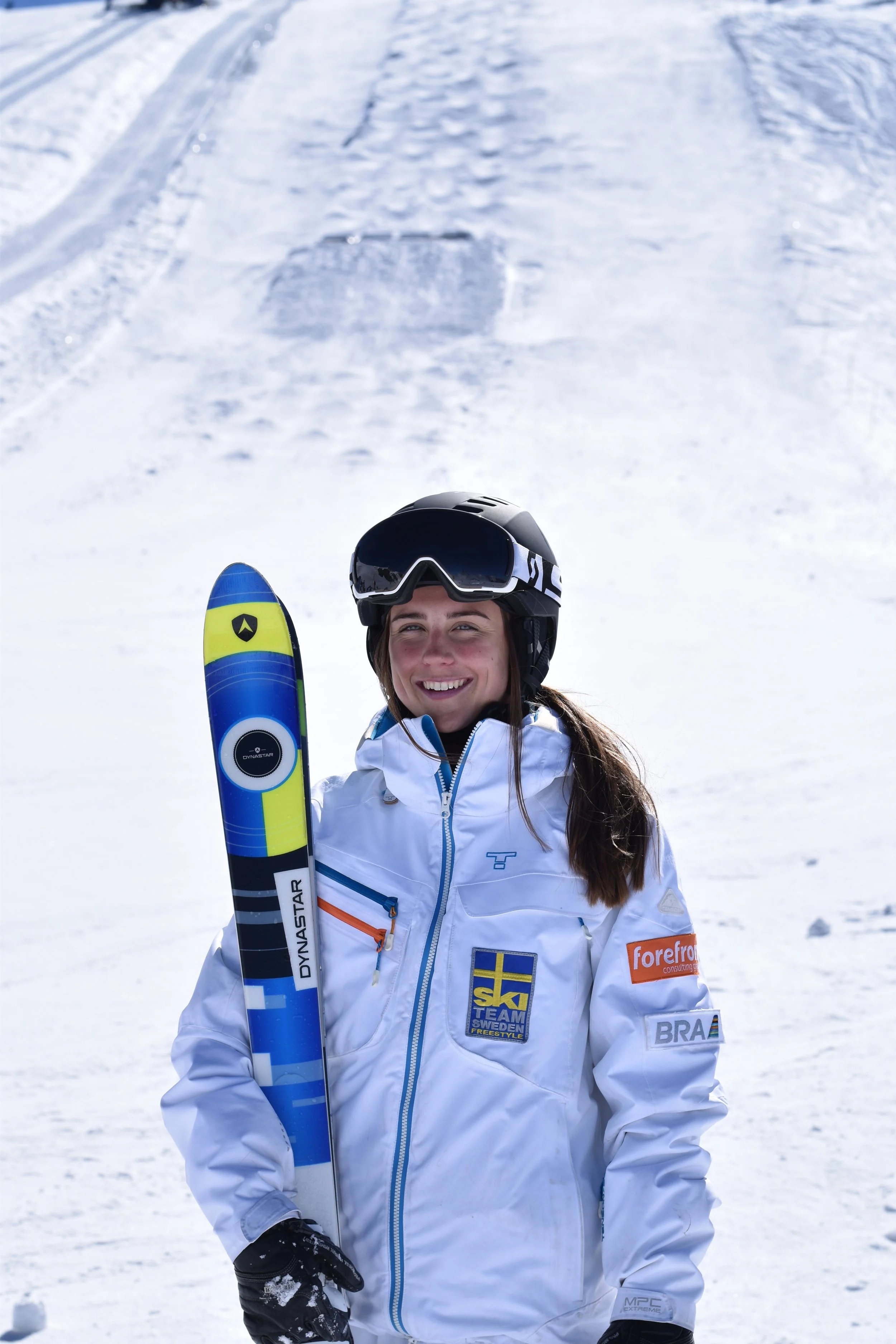 A female skier in a white jacket holding a colorful ski, standing on snow with a snowy ski slope in the background, smiling at the camera.