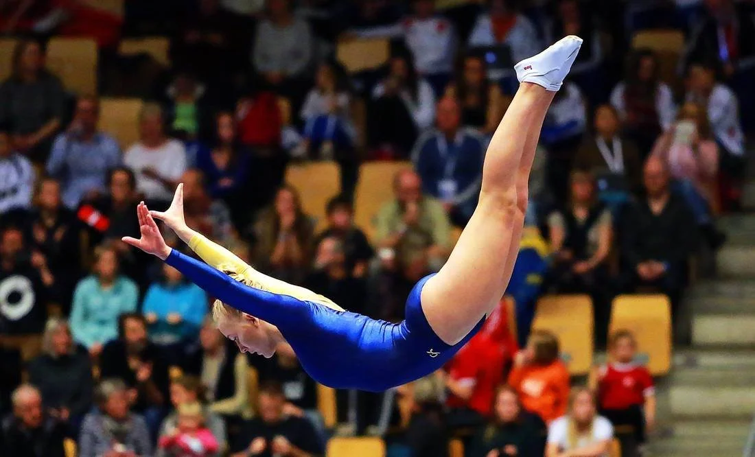 A female gymnast in a blue and yellow leotard performing a mid-air move during a gymnastics competition, with an audience watching in the background.
