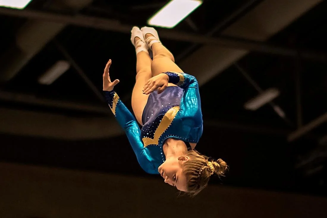 A female gymnast in a blue and gold leotard performing a flip in mid-air during a gymnastics routine.