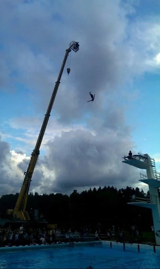 A person in mid-air jumping from a diving board into a swimming pool, with a crane and a person in the basket of the crane in the background under a cloudy sky, and a crowd watching.