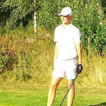A young person standing outdoors on a golf course, holding a golf club and wearing a white cap and white outfit.