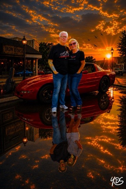 A couple standing in front of a red sports car during a sunset with a dramatic sky, reflections on the wet surface showing their reflection.