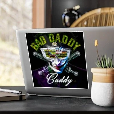 Digital tablet displaying a Joker face with the text 'Bad Daddy Caddy' and images of dumbbells and gym equipment, placed on a desk with a notebook, pen, and potted plant nearby.