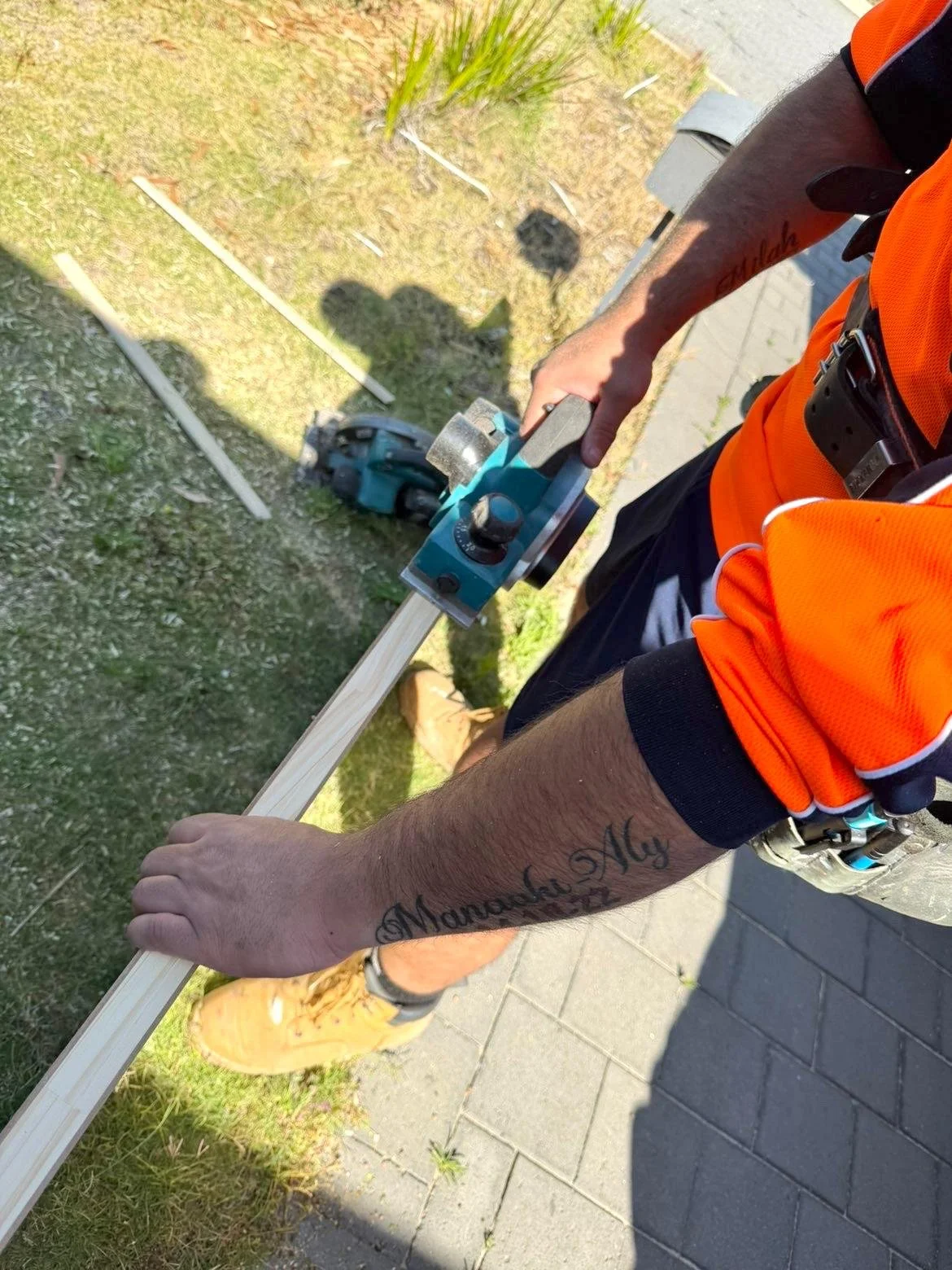 Person using a power tool to cut a wooden board outdoors on a grassy area near a sidewalk, wearing an orange safety vest and tan work boots.