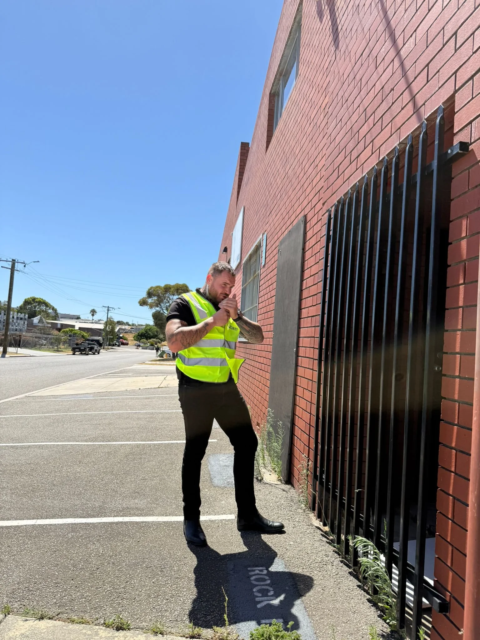 A man in a yellow safety vest standing outside a red brick building, lighting a cigarette near a black metal gate, with a clear blue sky in the background.