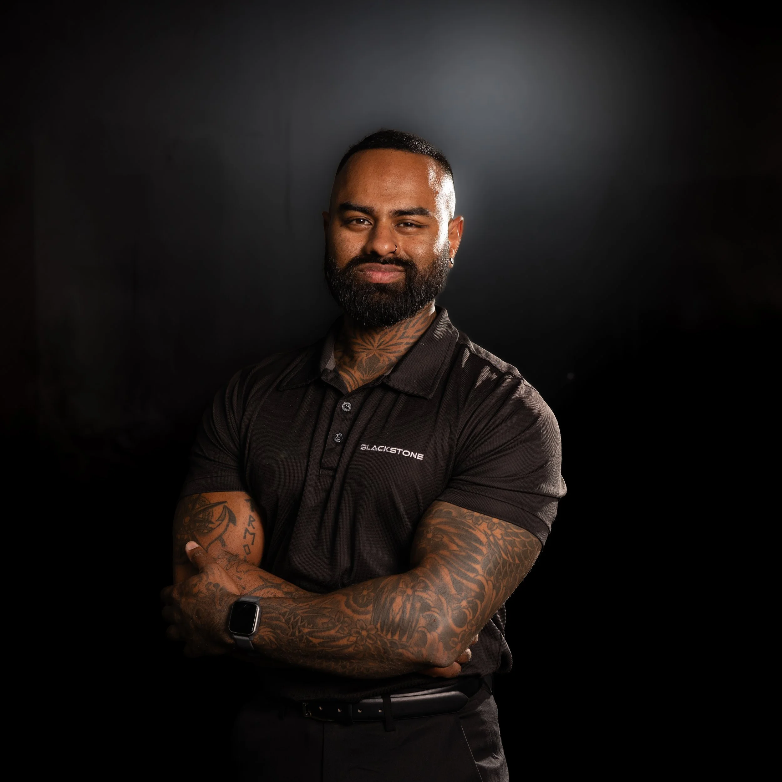 A man with a beard and tattoos on his arms, standing against a wooden wall, wearing a navy blue polo shirt with a logo that reads 'Blackstone'.