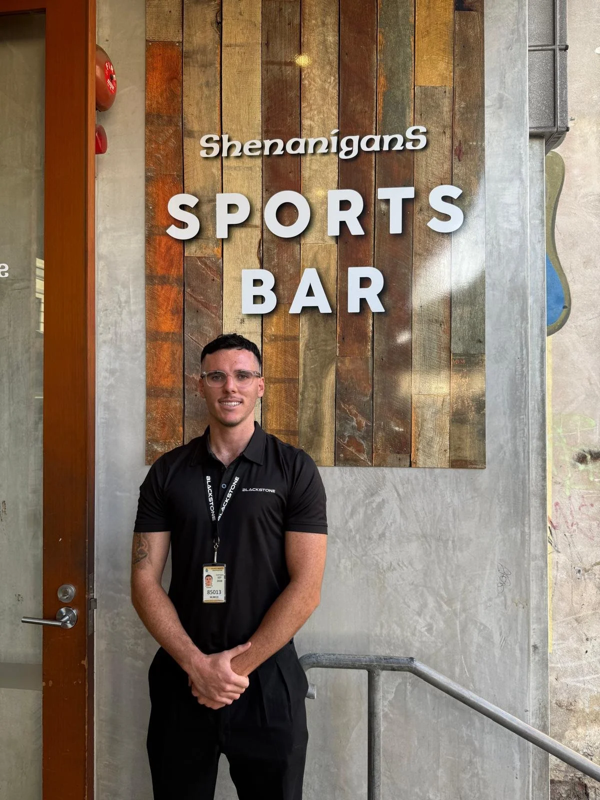 A young man with glasses, wearing a Blackstone uniform and a lanyard, standing in front of a sign that says 'ShenaniganS SPORTS BAR' in a restaurant or bar.