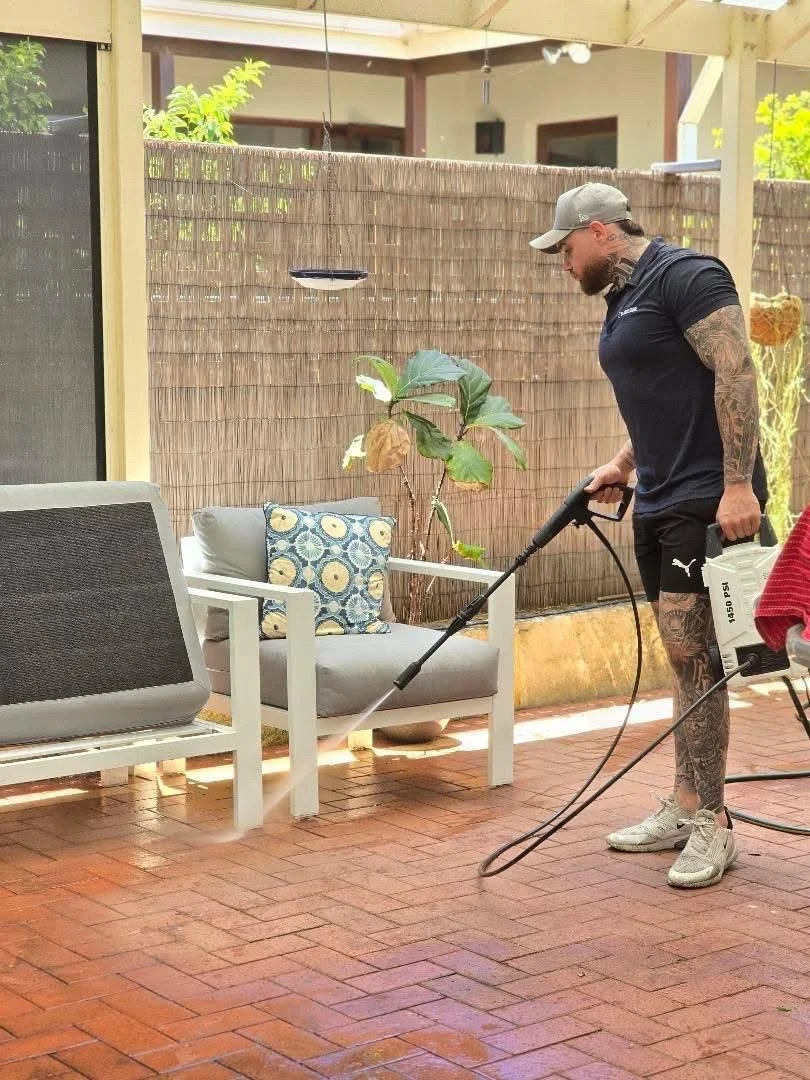 A man with tattoos, wearing a black Puma t-shirt, gray cap, and white sneakers, is pressure washing a patio with a pressure washer. The patio has brick flooring, outdoor furniture, and a small tree in a pot. A woven privacy fence and some greenery are in the background.