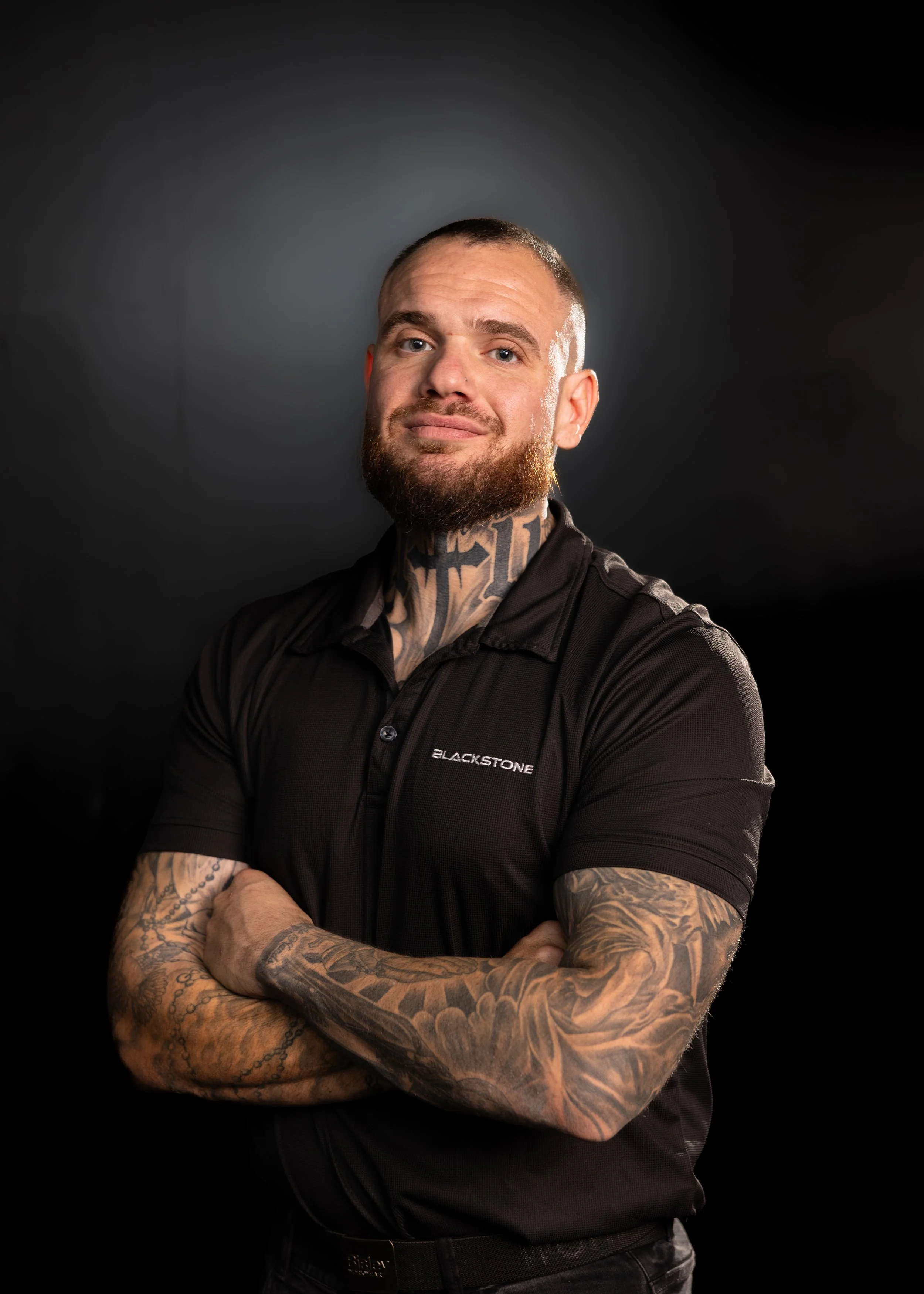 A man with tattoos on his arms and neck, wearing a black polo shirt with a logo, standing with arms crossed in front of a wooden wall, smiling at the camera.