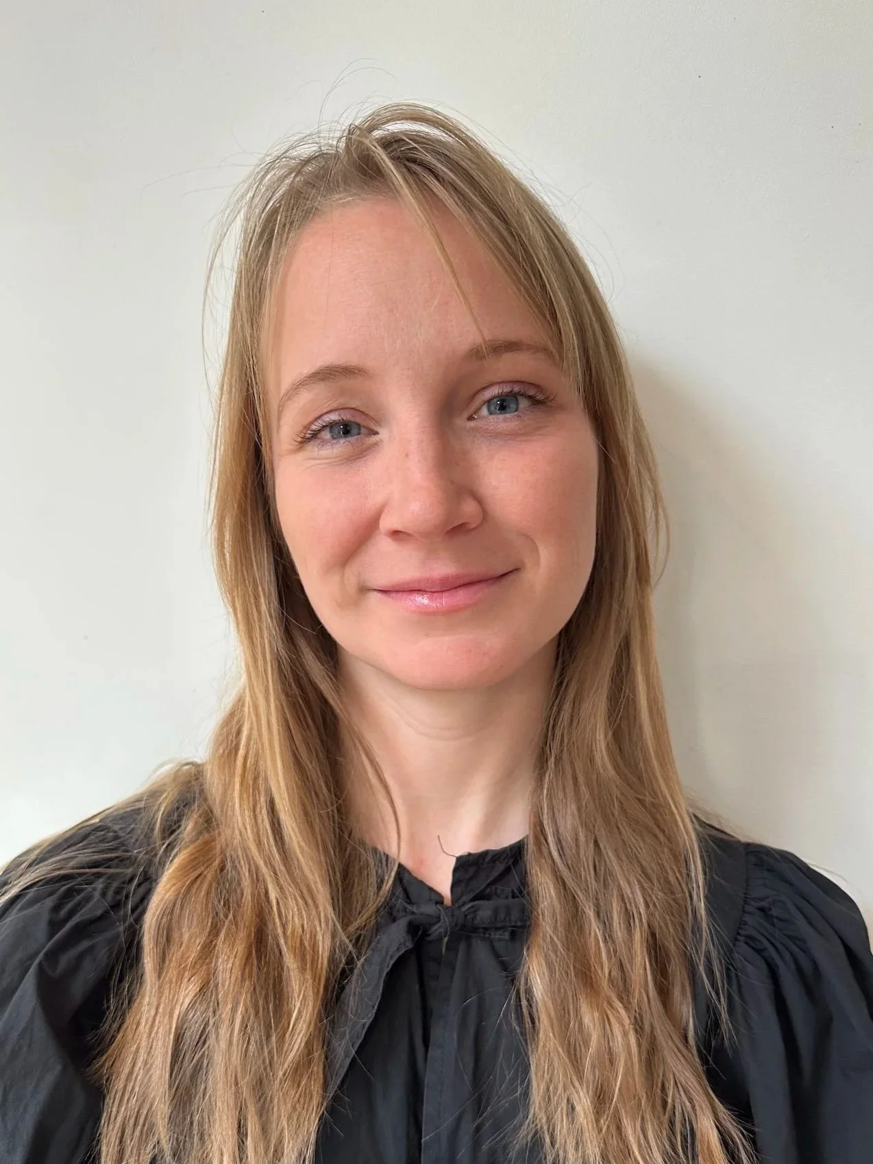 Rio Richardson Close-up of a woman with long blonde hair, light skin, and blue eyes smiling, standing against a plain white wall.