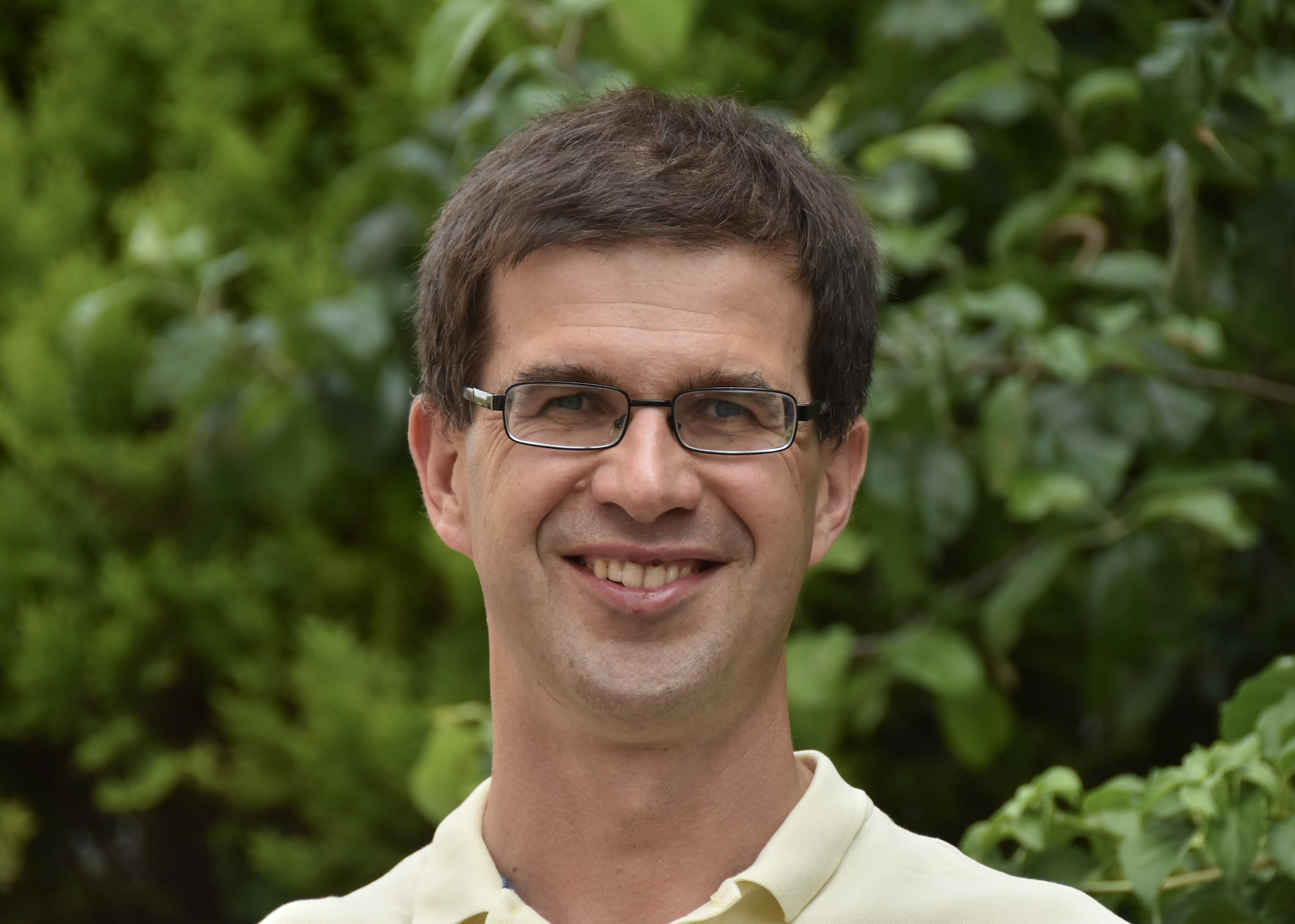 Oleksandr Kit Smiling man with glasses outdoors, surrounded by green foliage.