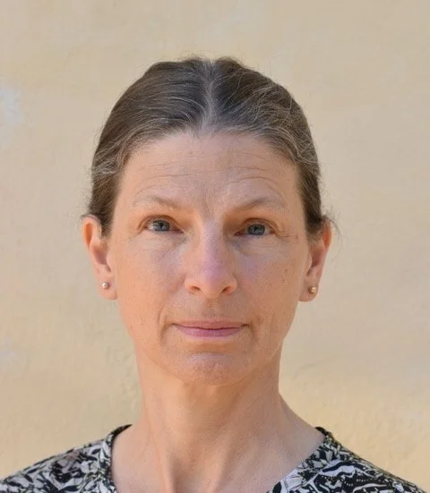 Claire Wigg Close-up portrait of a mature woman with light skin, brown hair tied back, wearing pearl earrings and a black and white patterned top, standing against a beige wall.