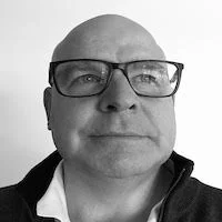 Nick Gogerty Close-up of a middle-aged man with glasses, bald head, wearing a collared shirt, smiling slightly in black and white.
