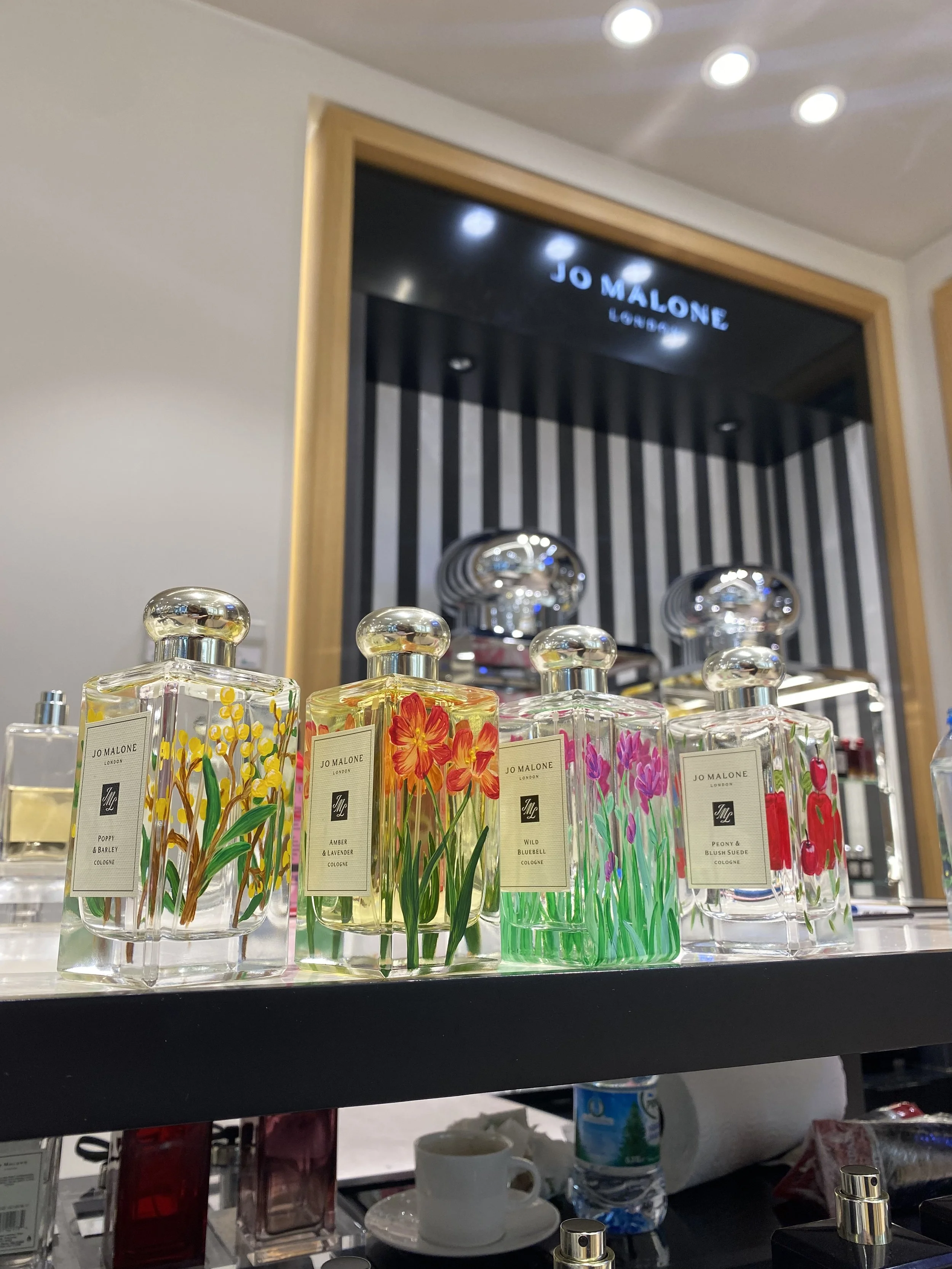 Display of Jo Malone London perfume bottles with floral designs, including poppy, amber, violet, peony, and bluebell scents, on a retail counter in a store.