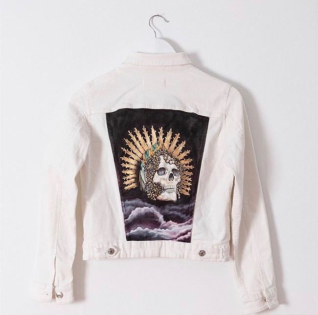 White denim jacket with a large embroidered patch on the back featuring a skull with a crown of golden spikes and feathers, set against dark clouds.