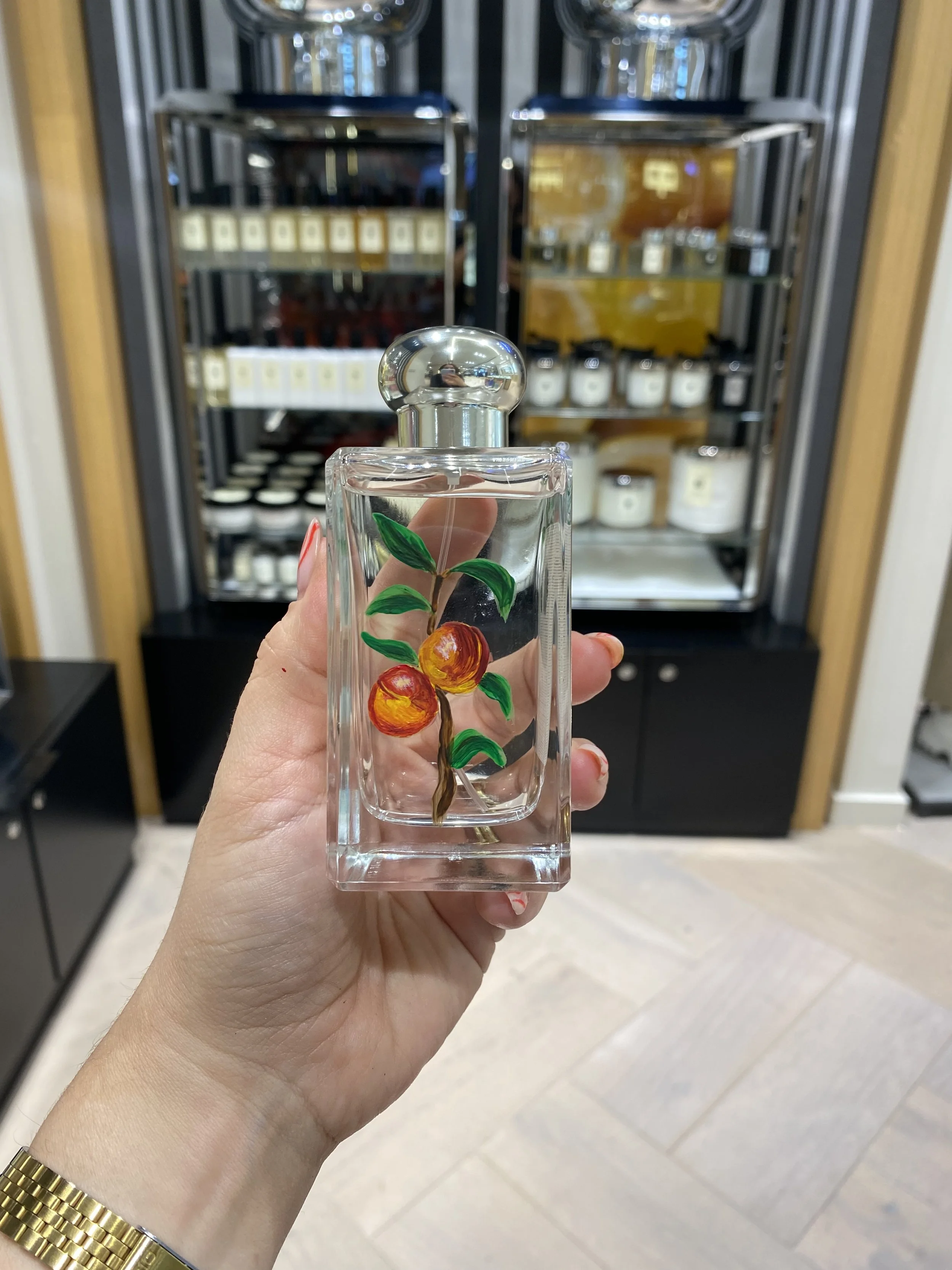 A hand holding a glass perfume bottle with painted peach or apricot fruit and green leaves, in front of a refrigerated display of jars and bottles.