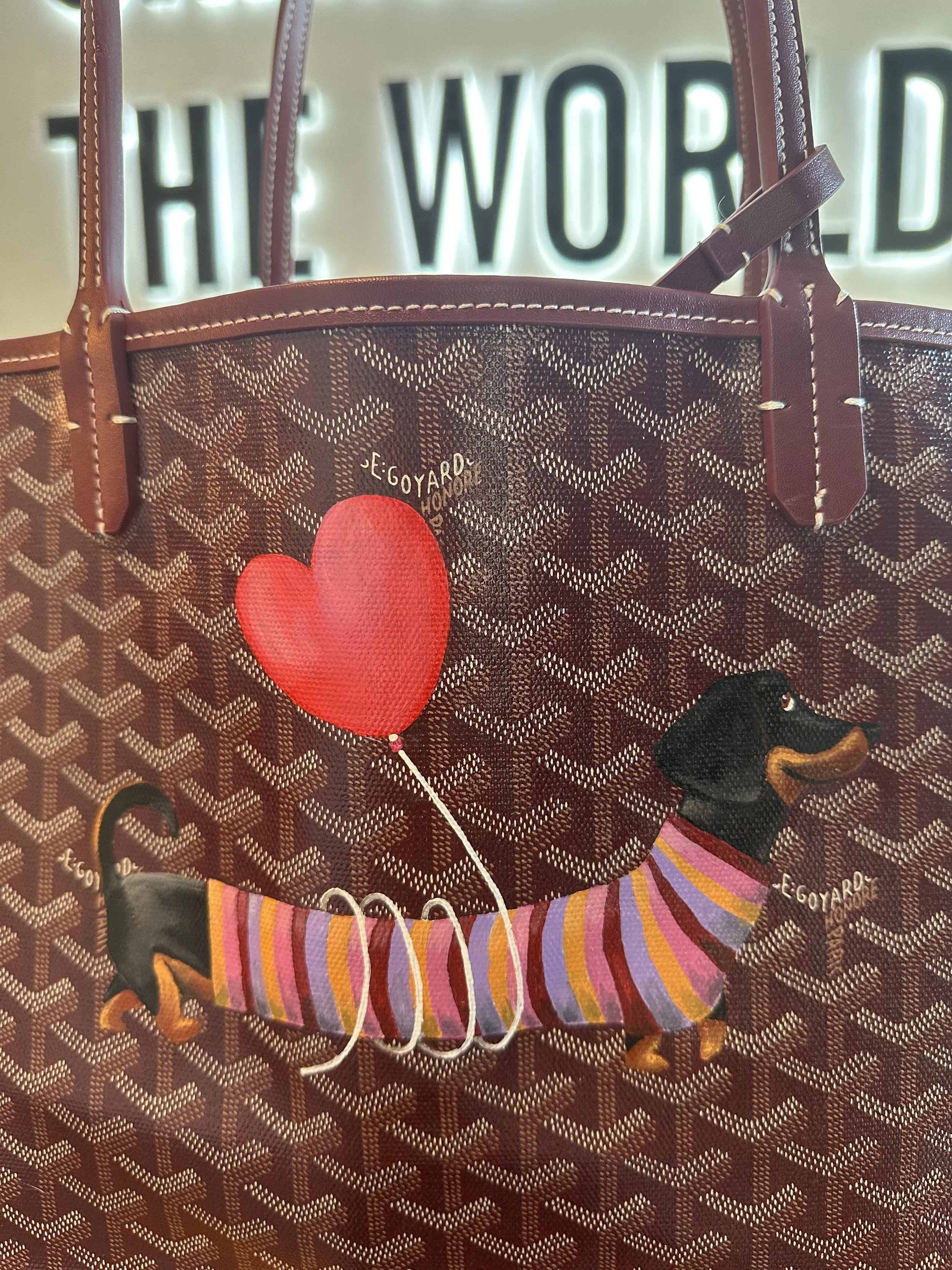 A patterned brown tote bag with a design of a dachshund wearing a striped purple, pink, and yellow sweater, holding a red heart-shaped balloon. The background has large black and white lettering that says 'THE WORLD'.