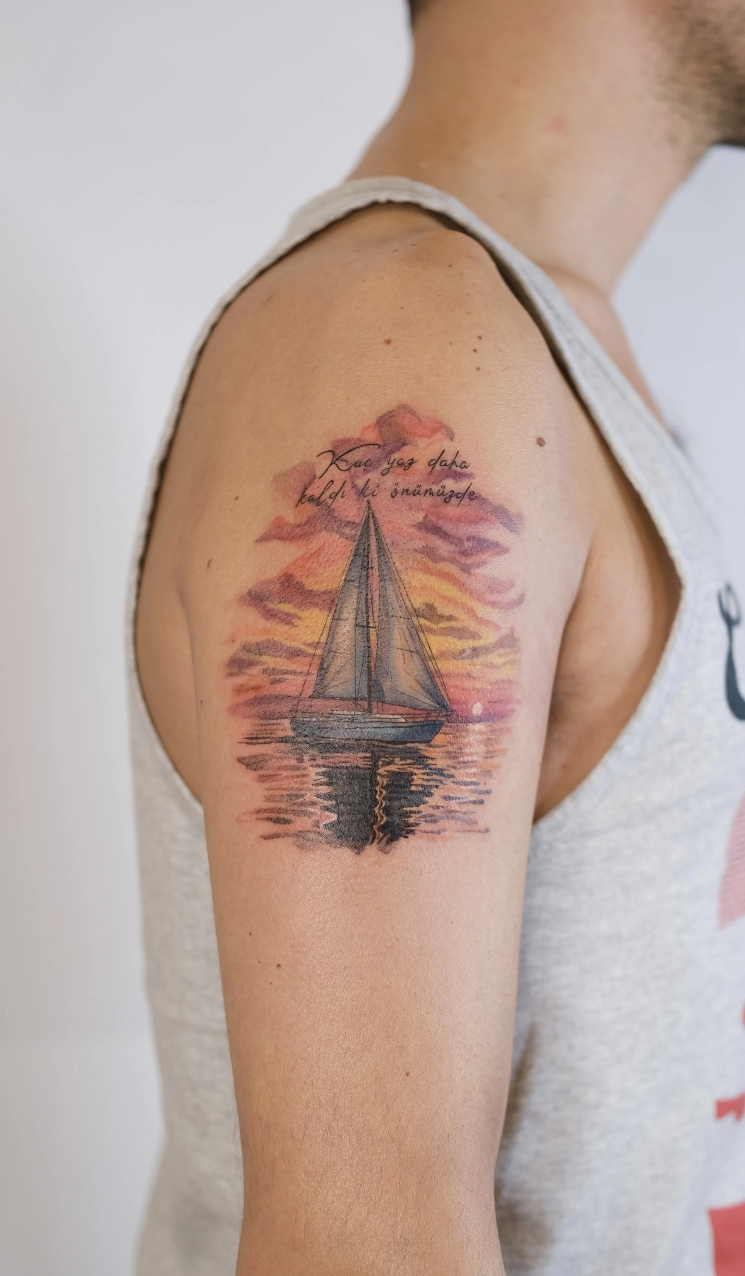 Tattoo of a sailboat on the upper arm, with a sunset and water reflection, including a quote in Turkish.