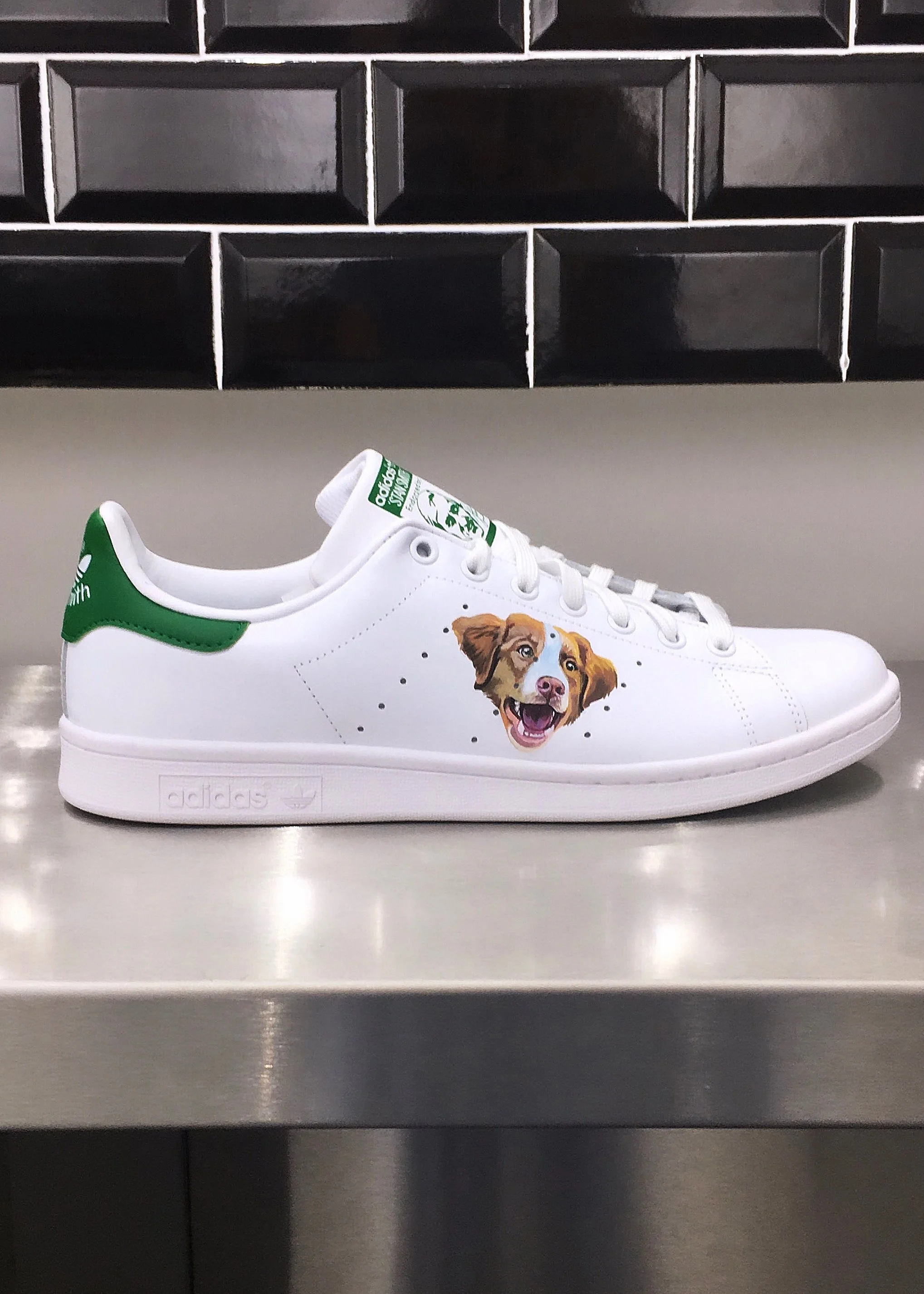 A white sneaker with green accents and a colorful dog face graphic on the side, placed on a metallic surface.