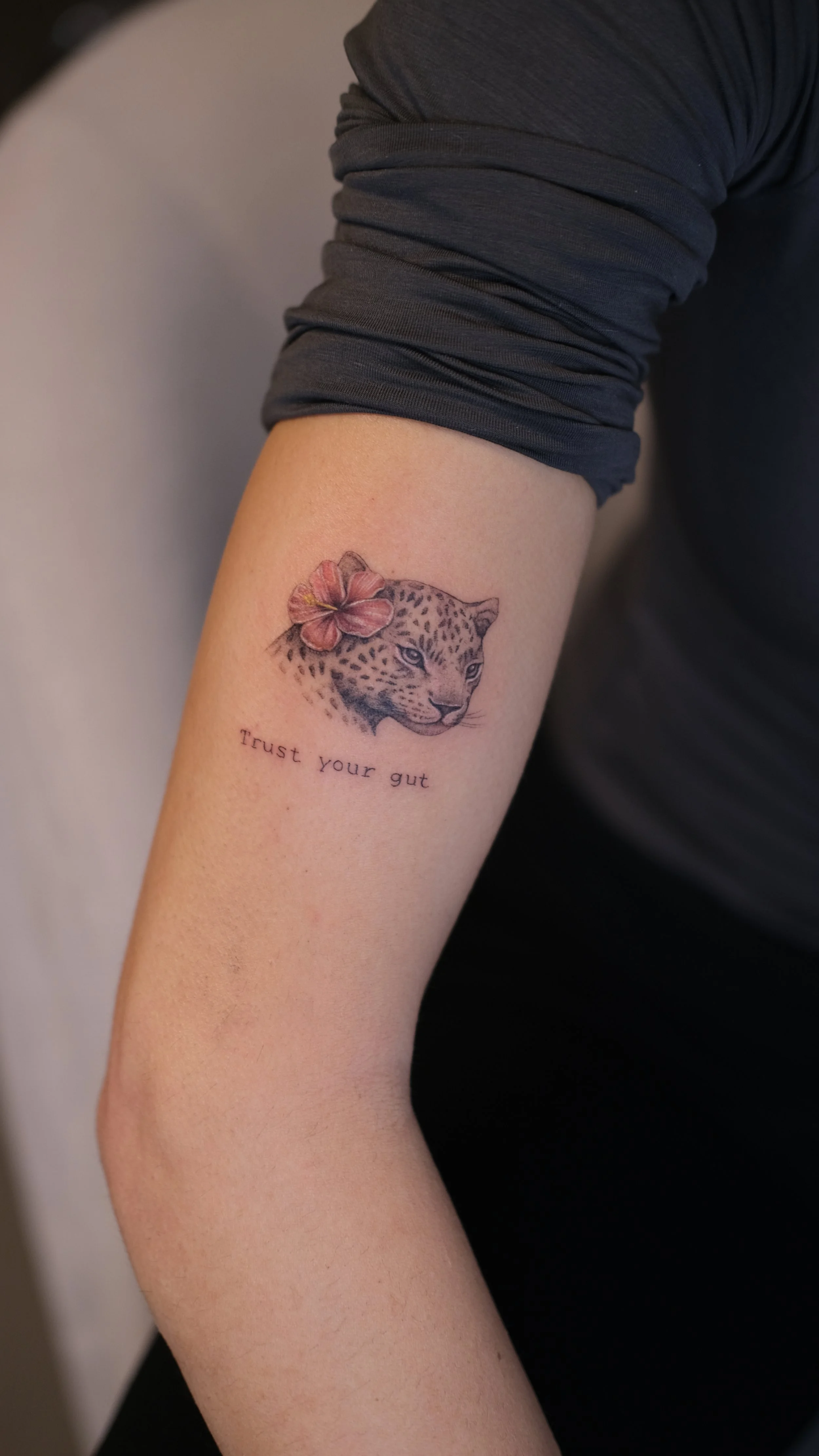 Tattoo of a leopard's face with a pink flower on its head and the words 'Trust your gut' below.
