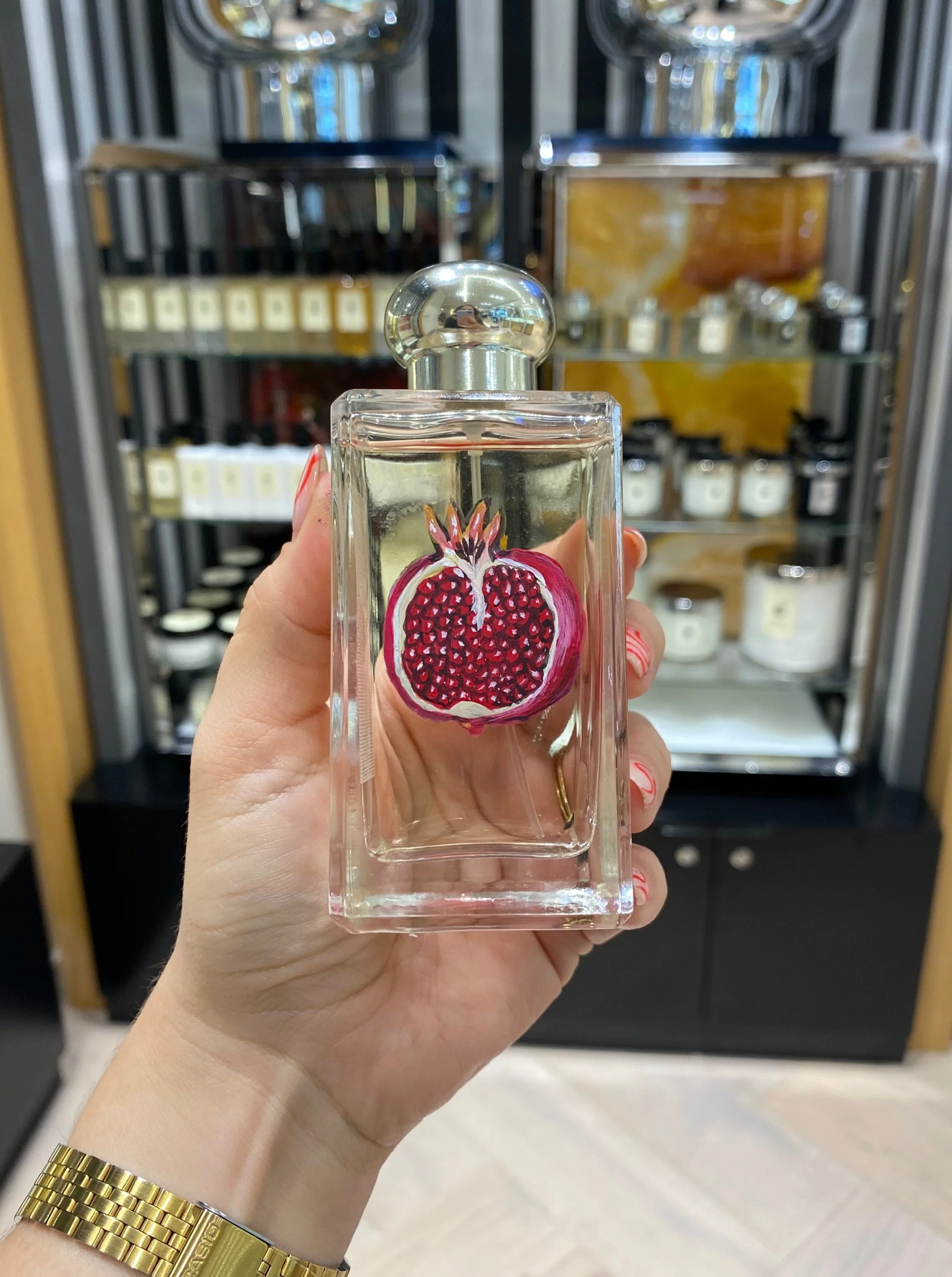 Hand holding a glass perfume bottle with pomegranate illustration, against a background of spice jars or perfumes on shelves.