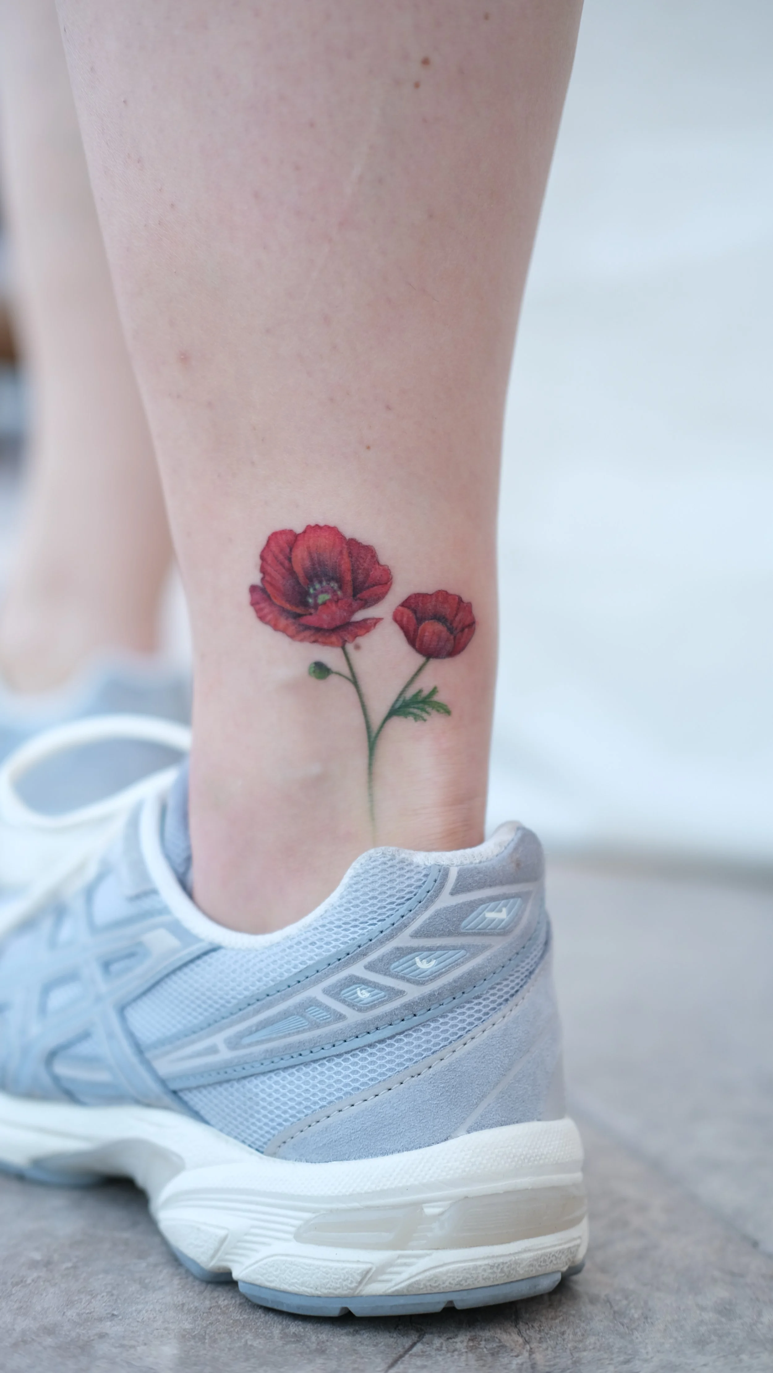 A person's leg with a tattoo of two red poppy flowers, wearing a white sneaker on a concrete floor.