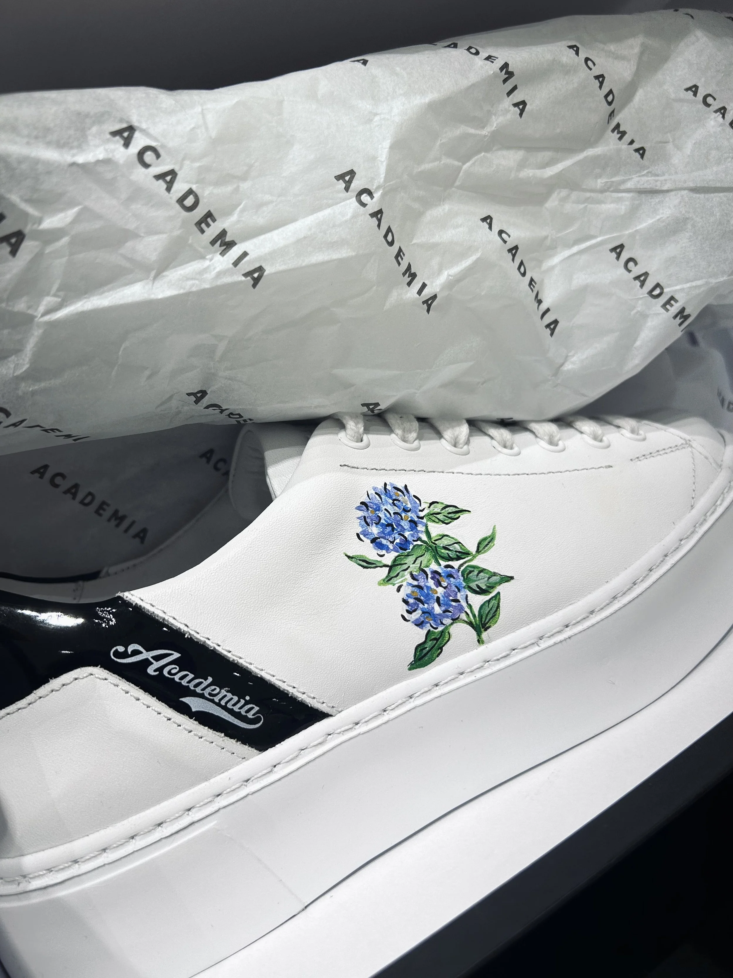 White sneaker with floral embroidery on the side, labeled 'Academia,' inside a box with tissue paper printed with the word 'ACADEMIA' repeatedly.