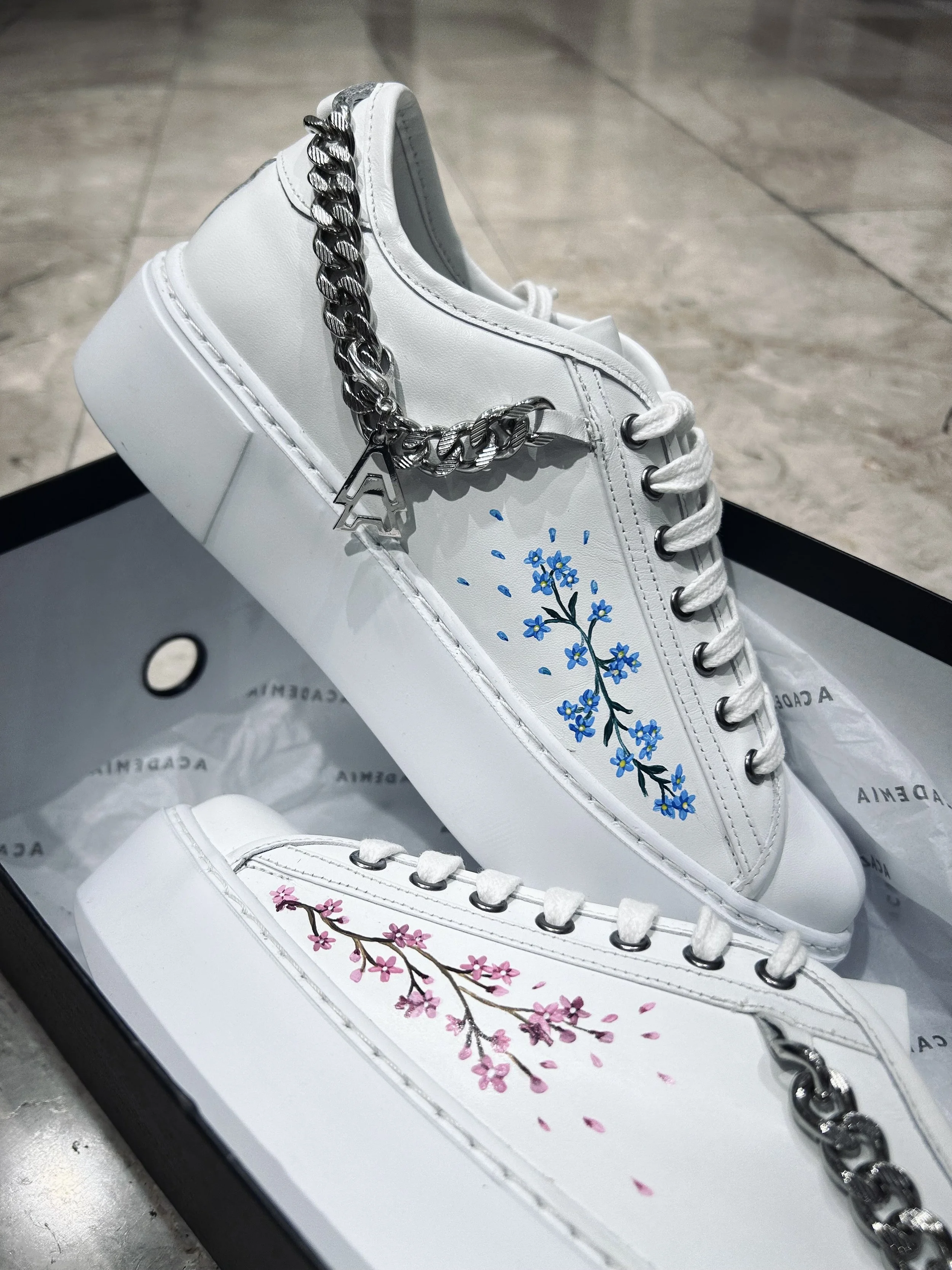 Pair of white sneakers with painted blue and pink flower designs, each featuring a silver chain with a letter 'A' charm draped over the shoes, placed inside a black box with tissue paper.