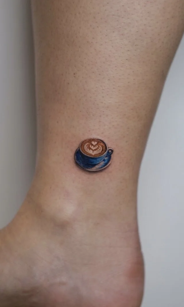 A small tattoo of a blue coffee cup with latte art on a person's ankle.