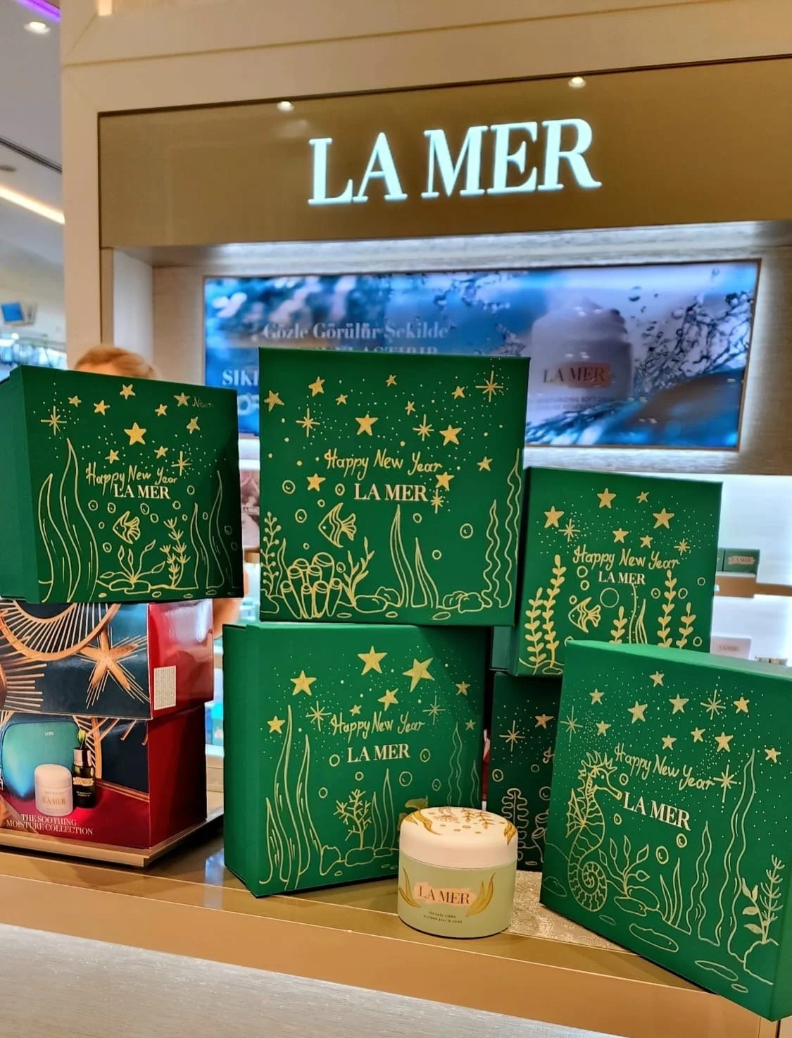 Holiday gift sets of La Mer skincare products in green boxes with star and seaweed designs, and a small jar of La Mer cream in front, on a retail display.