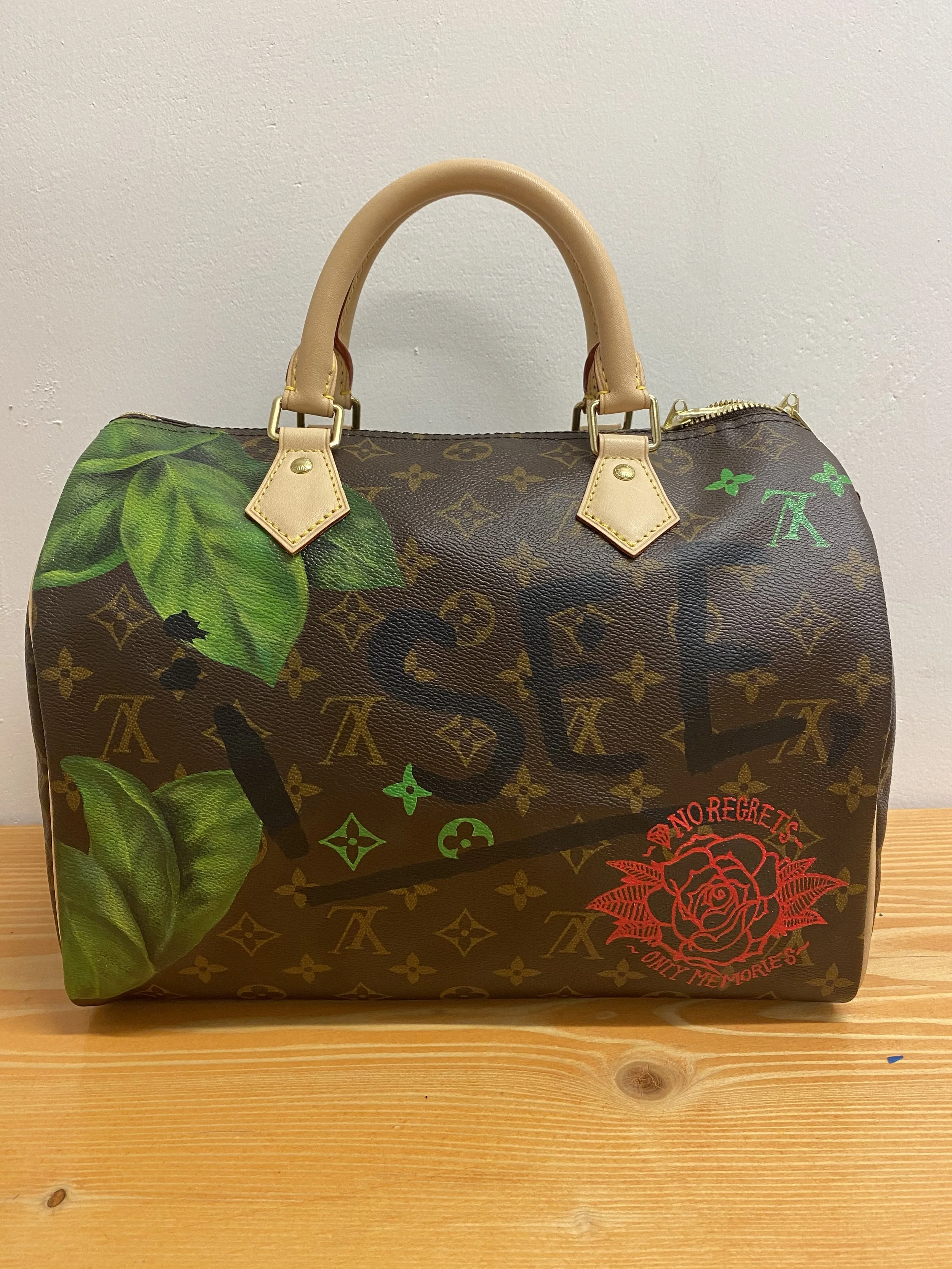 A designer handbag with a brown monogram pattern, green leaf prints, red text that says 'No Regrets', and a red flower graphic, placed on a wooden surface against a plain wall.