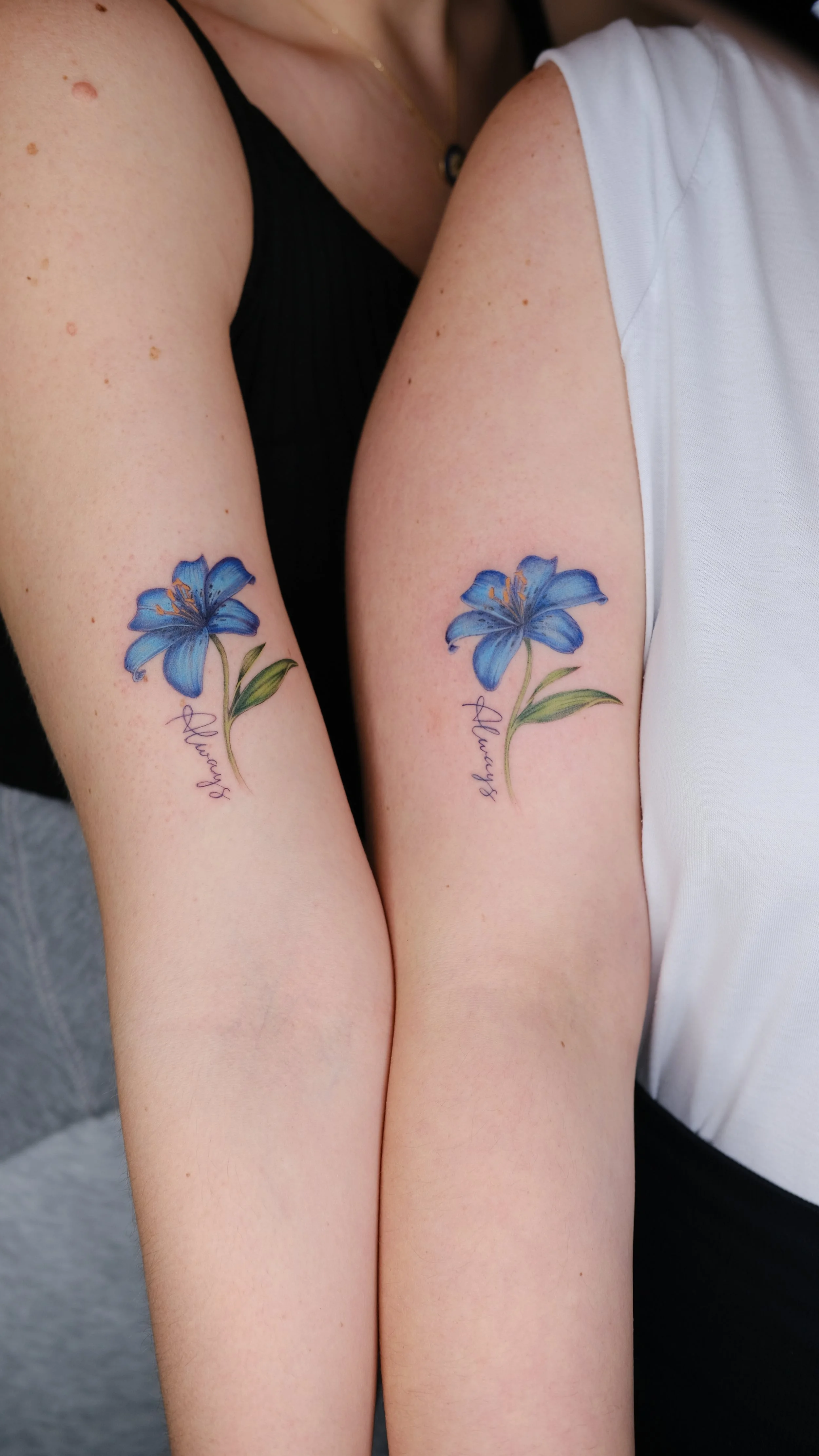 Two people with matching blue lily tattoos on their forearms, with the word 'Always' written below each flower.