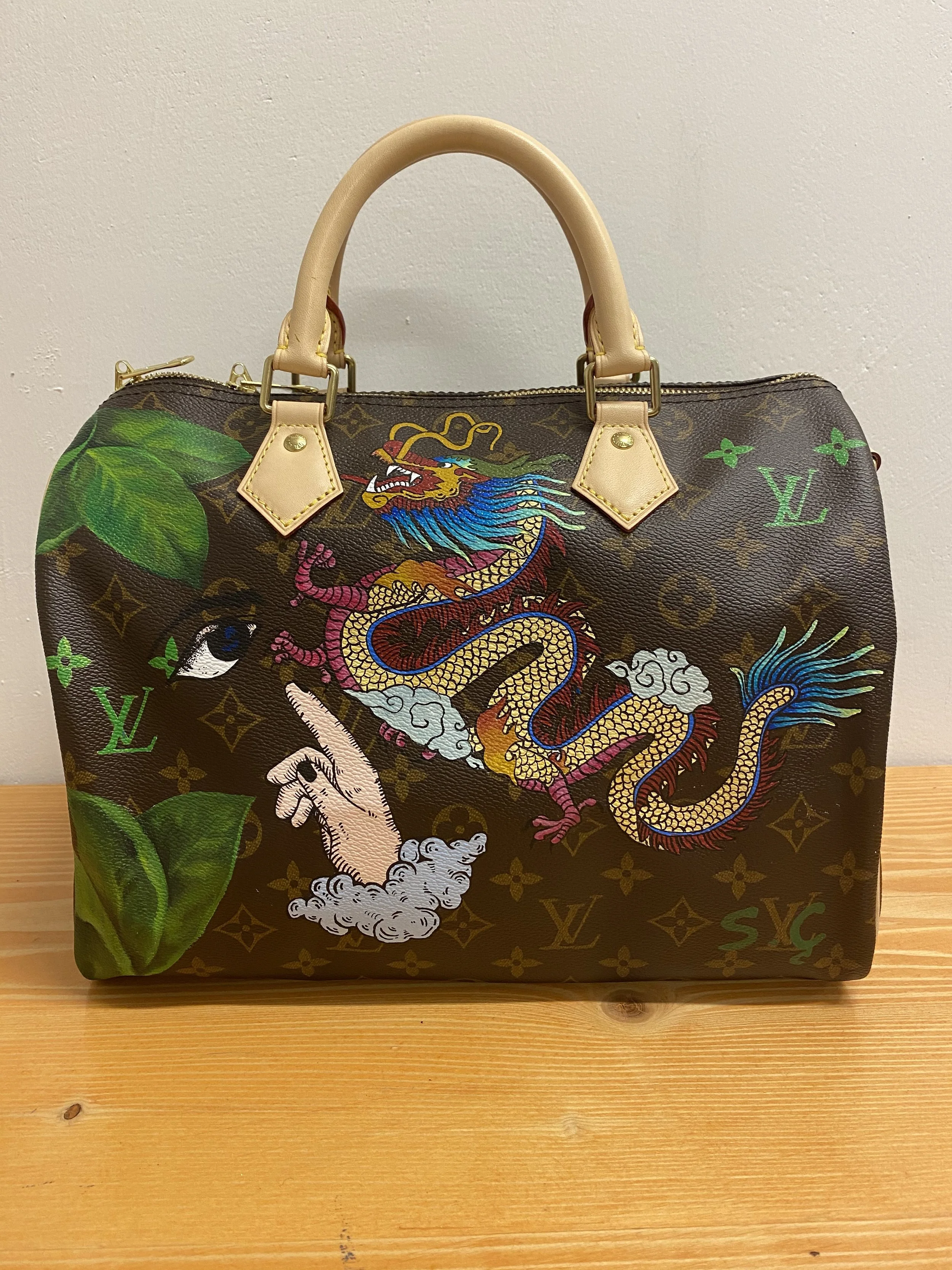 A Louis Vuitton handbag with a colorful dragon and mystical illustrations, including clouds and a pointing hand, on a brown monogram canvas.