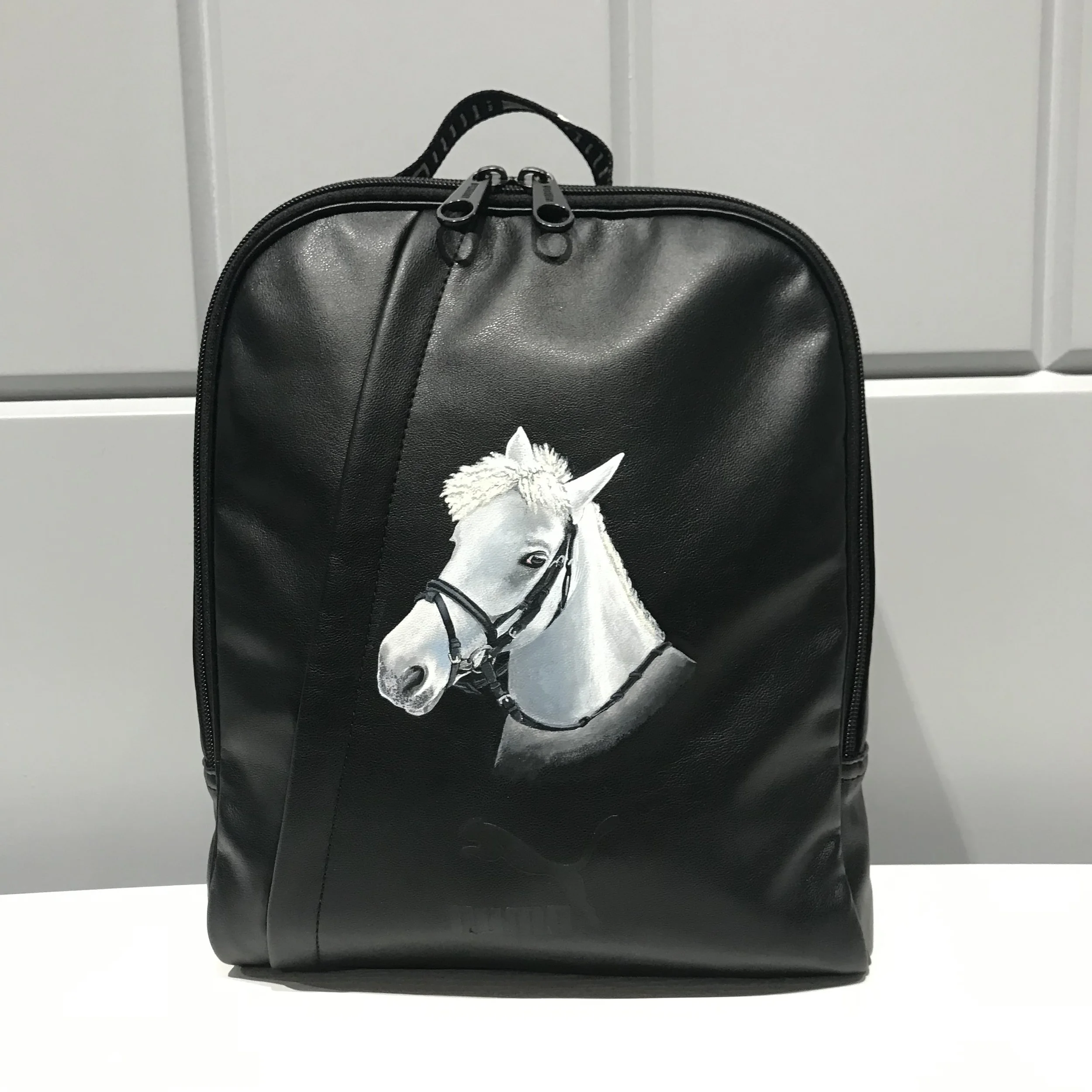 A black backpack with a white horse head painted on the front, featuring a bridle and reins.