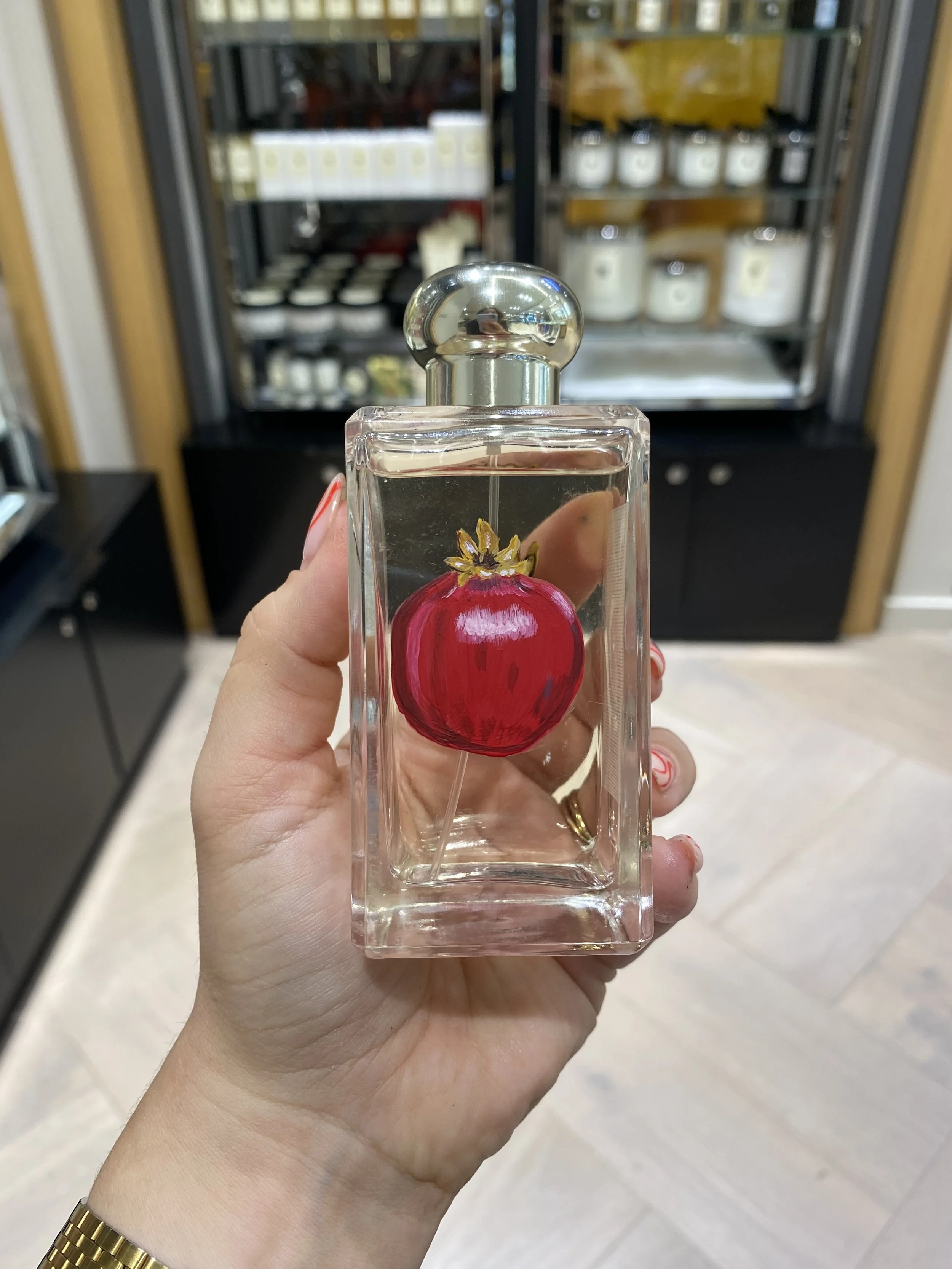 Hand holding a glass perfume bottle with a painted pomegranate on it, in front of a refrigerated display case.