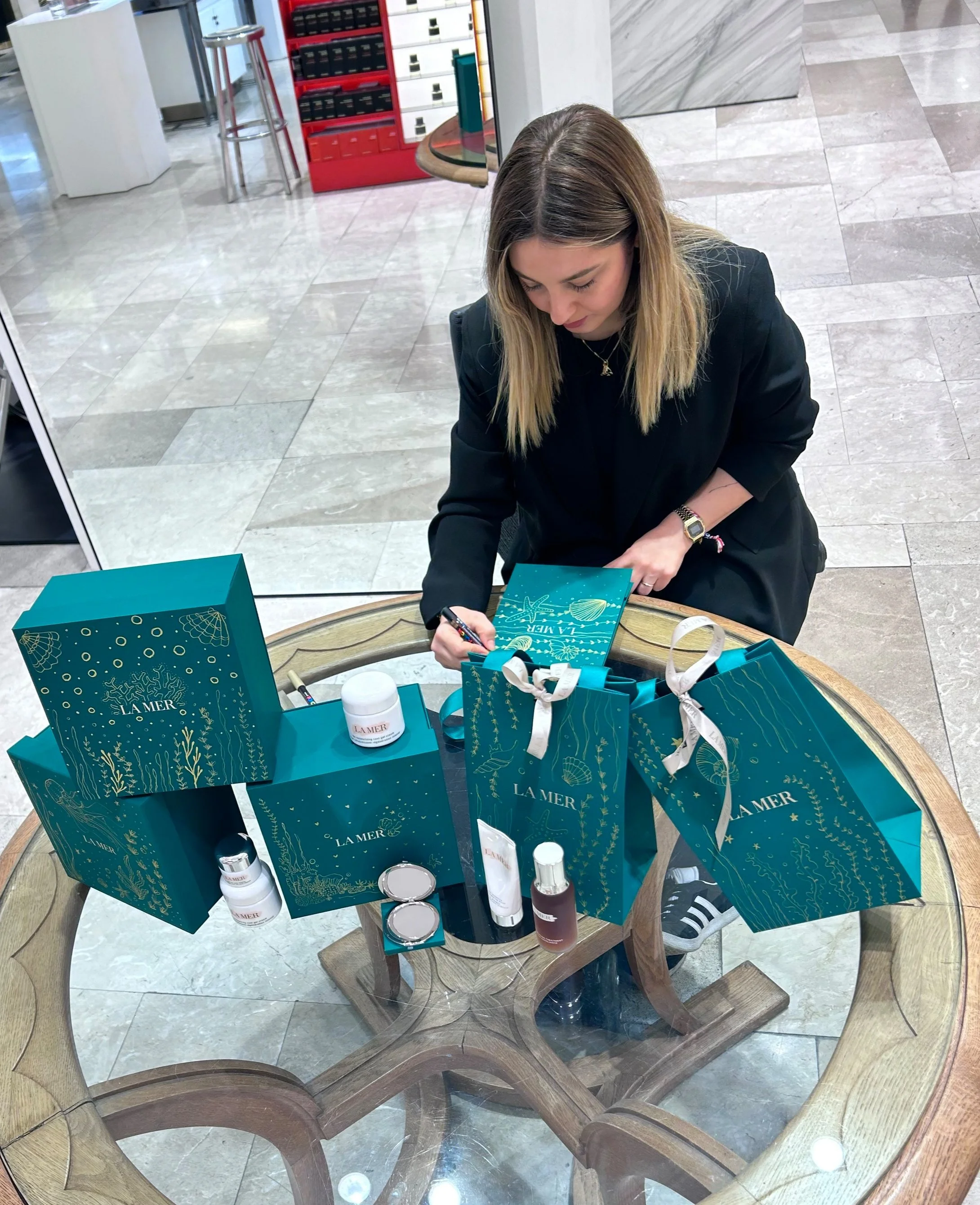 A woman painting an item at a display of La Mer skincare products in teal shopping bags. The products are arranged on a wooden table.