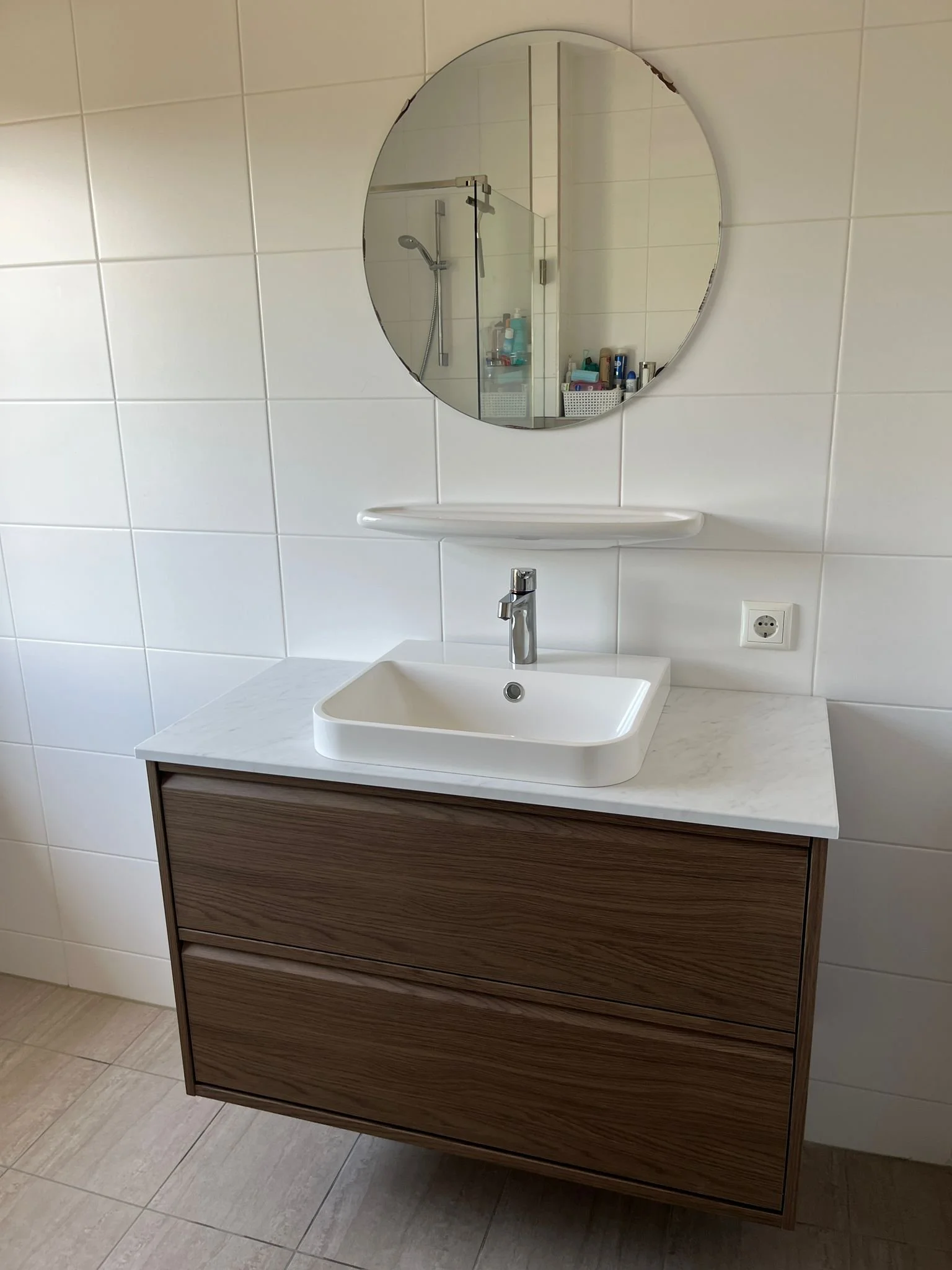 Witte wastafel met een houten kast eronder, een ronde spiegel, en een muur met witte tegels.