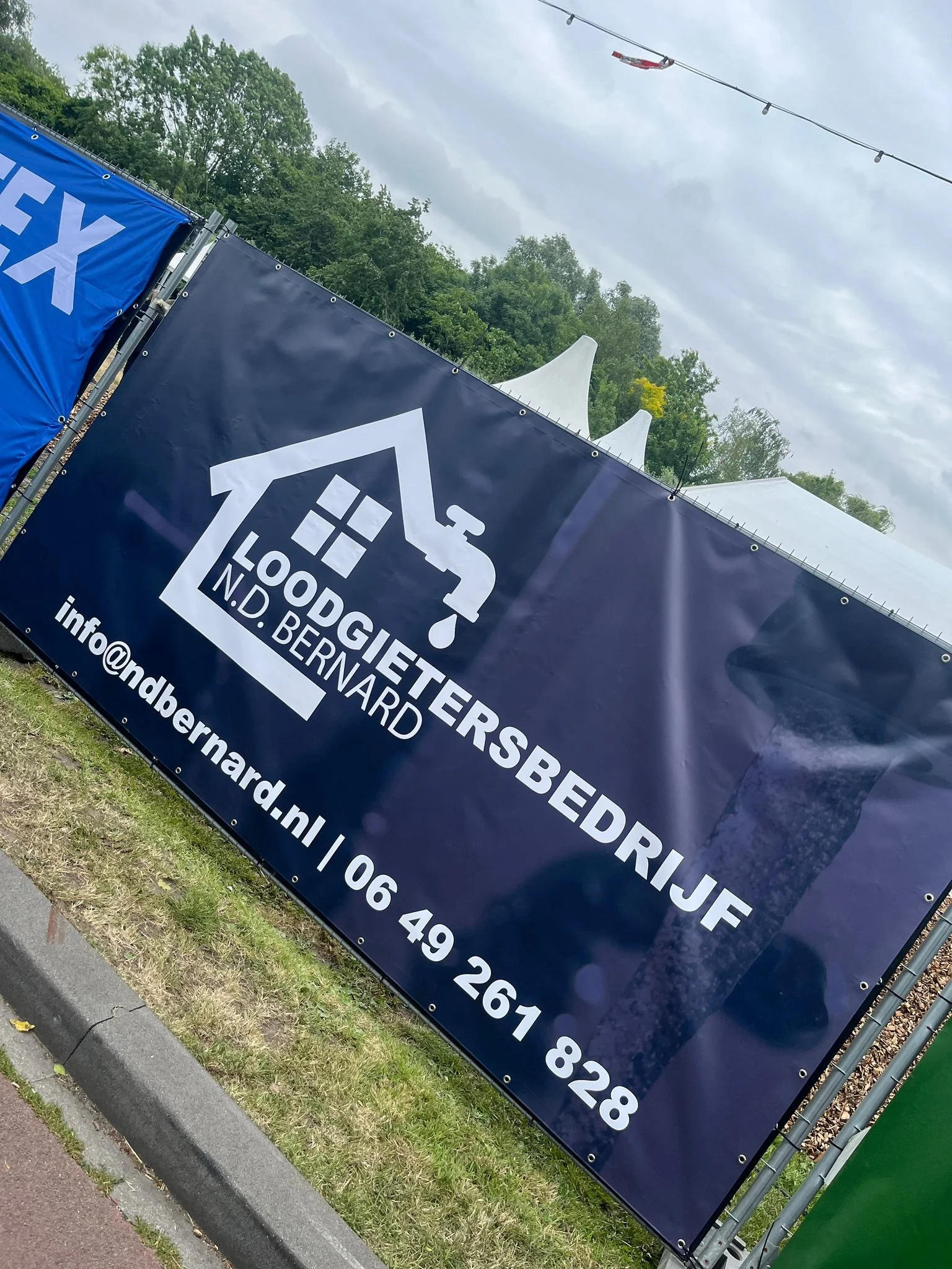 Een blauwe reclameborden voor loodgietersbedrijf N.D. Bernard, inclusief het logo van een waterkraan en contactinformatie onder een bewolkte hemel met groen omringde bomen op de achtergrond.