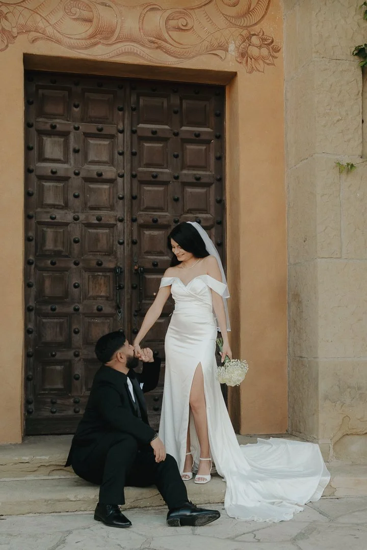 A bride and groom pose in front of a large wooden door, with the groom kneeling and kissing the bride's hand. The bride wears a white wedding gown with a slit, veil, and holds a bouquet of flowers. The groom is dressed in a black suit with a white sh