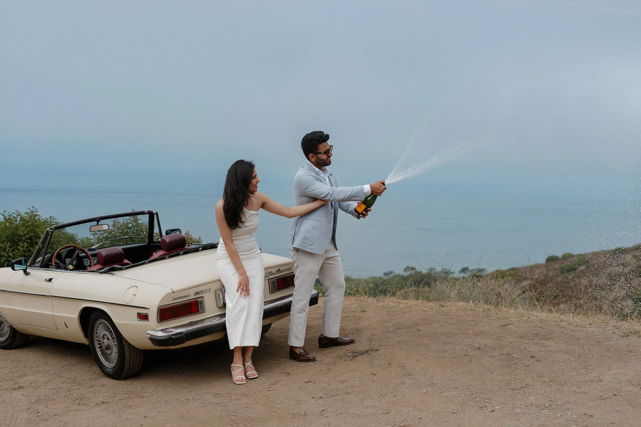A man in a light gray suit and a woman in a white strapless dress are standing next to a cream-colored convertible car, celebrating outdoors on a cloudy day. The man is opening a bottle of champagne, and the woman is smiling. The car is parked on a d