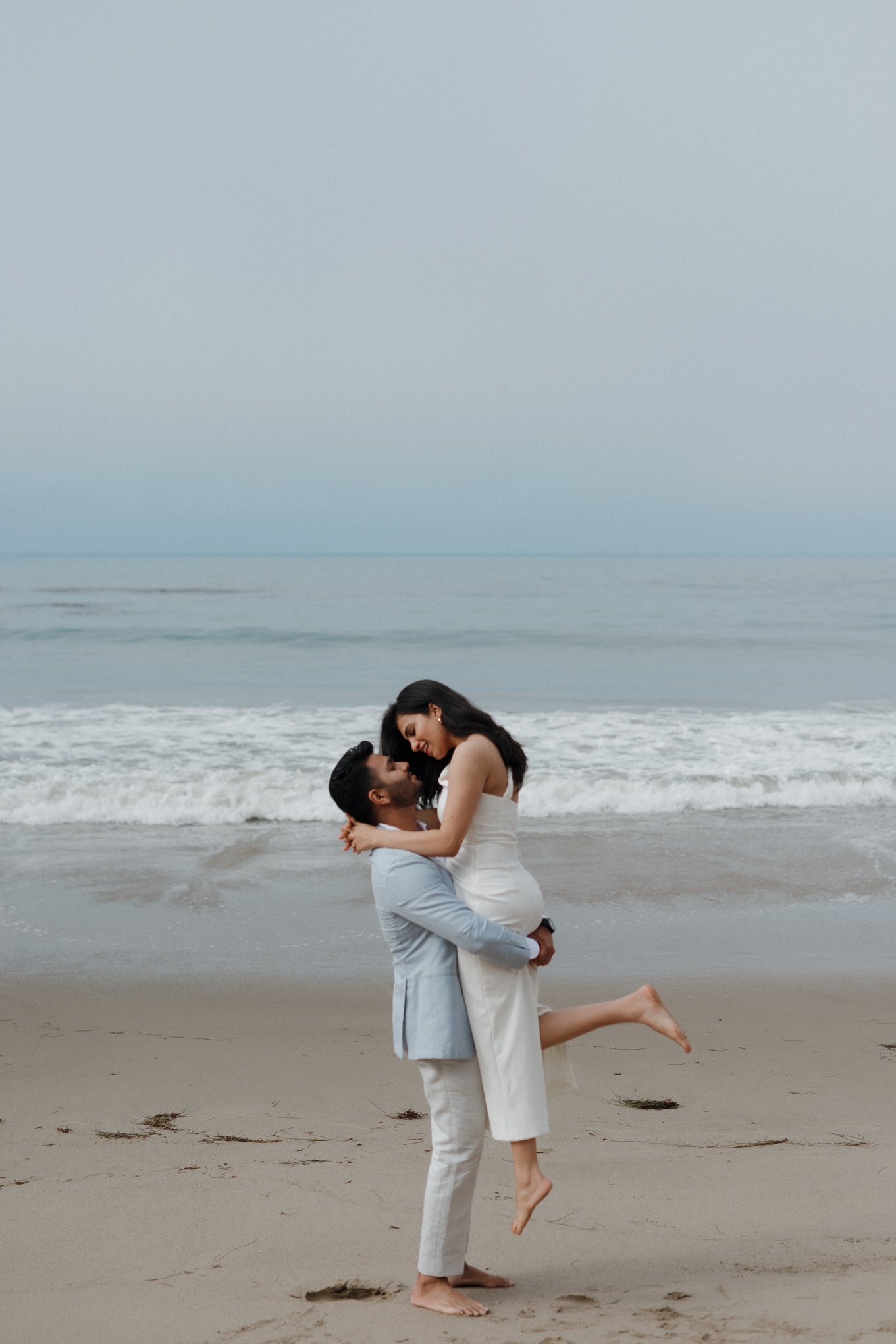 A couple on the beach with the man lifting the woman, both smiling, with the ocean and cloudy sky in the background.
