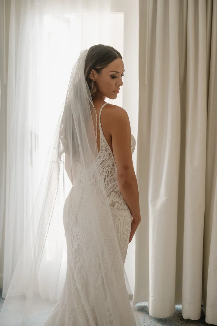 Bride in a lace wedding dress and veil standing by curtains