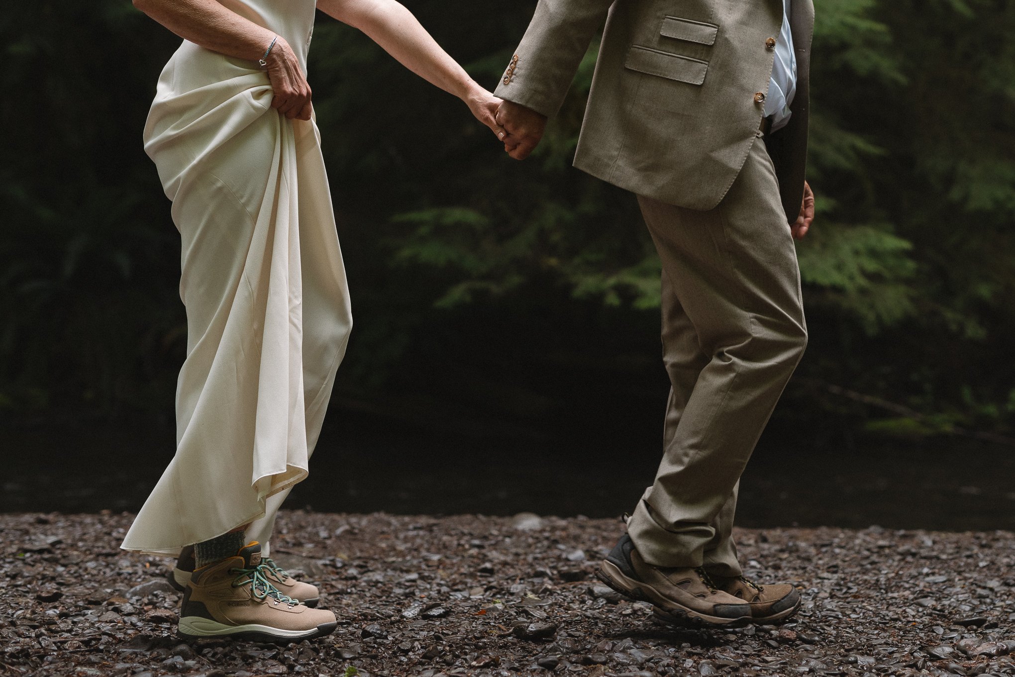 A bride and groom holding hands and standing on a gravel path outdoors. The bride is in a white dress and hiking boots, and the groom is in a tan suit and hiking boots.